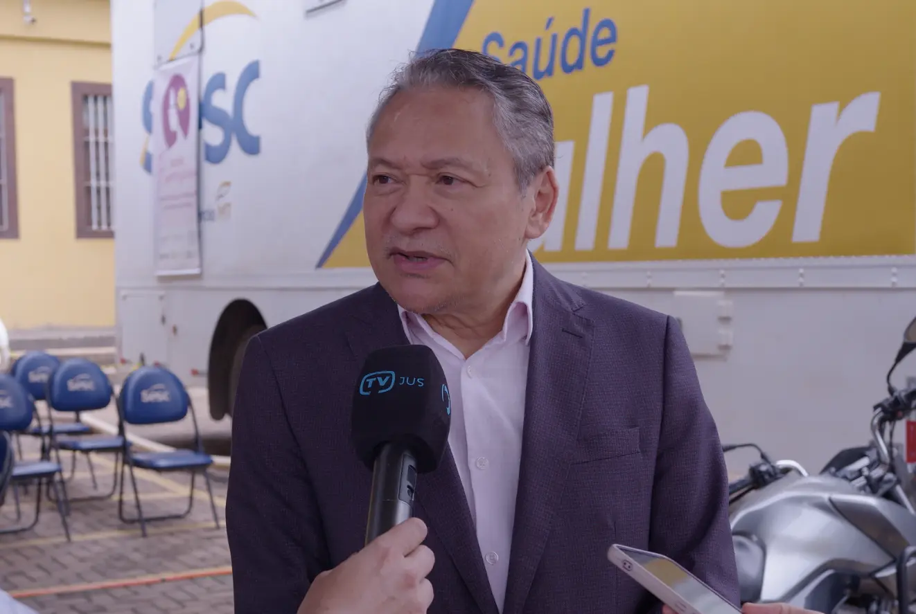 Desembargador Mário Kono durante entrevista à TV.Jus, em frente à carreta Sesc Saúde Mulher. Ele é um senhor pardo, de cabelos grisalhos, com traços orientais, usando camisa rosa clara e terno cinza.