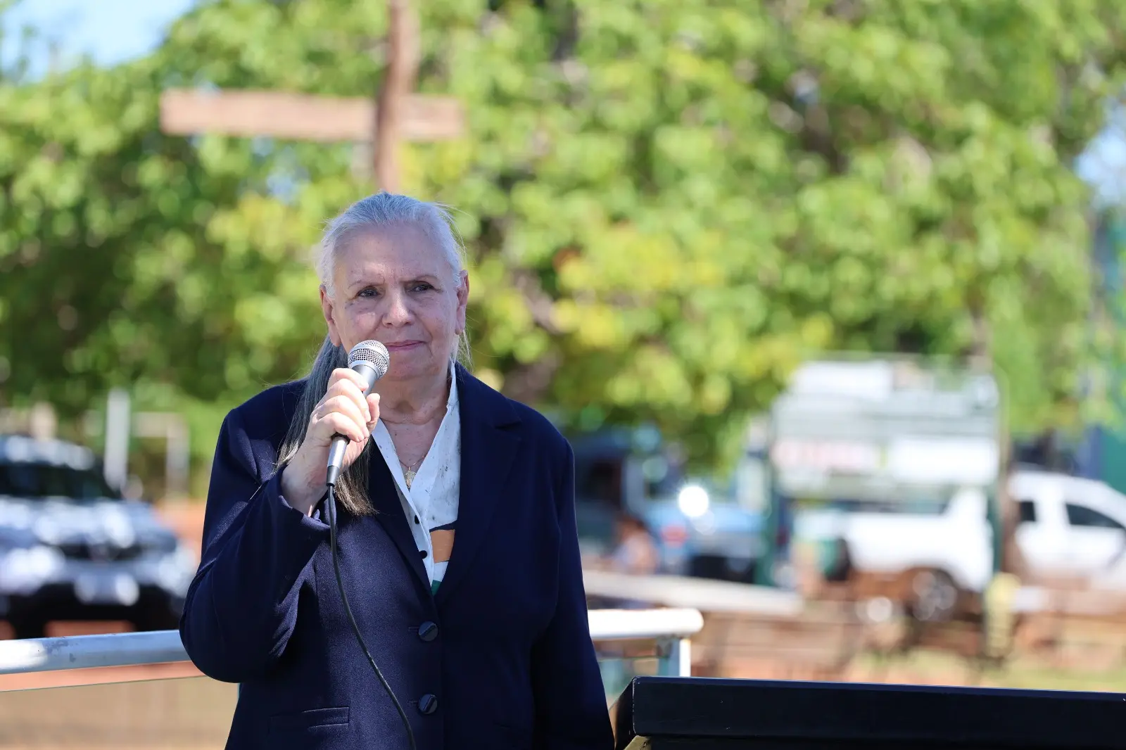 Mulher de cabelos brancos e terno azul escuro fala ao microfone em ambiente externo. Árvores verdes desfocadas aparecem ao fundo sob céu claro.