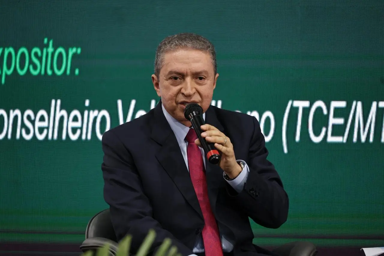 Conselheiro do Tribunal de Contas de Mato Grosso, Valter Albano, sentado e falando ao microfone. Ele é um senhor magro, pardo , de cabelos grisalhos, usando camisa azul, gravata vermelha e terno preto.