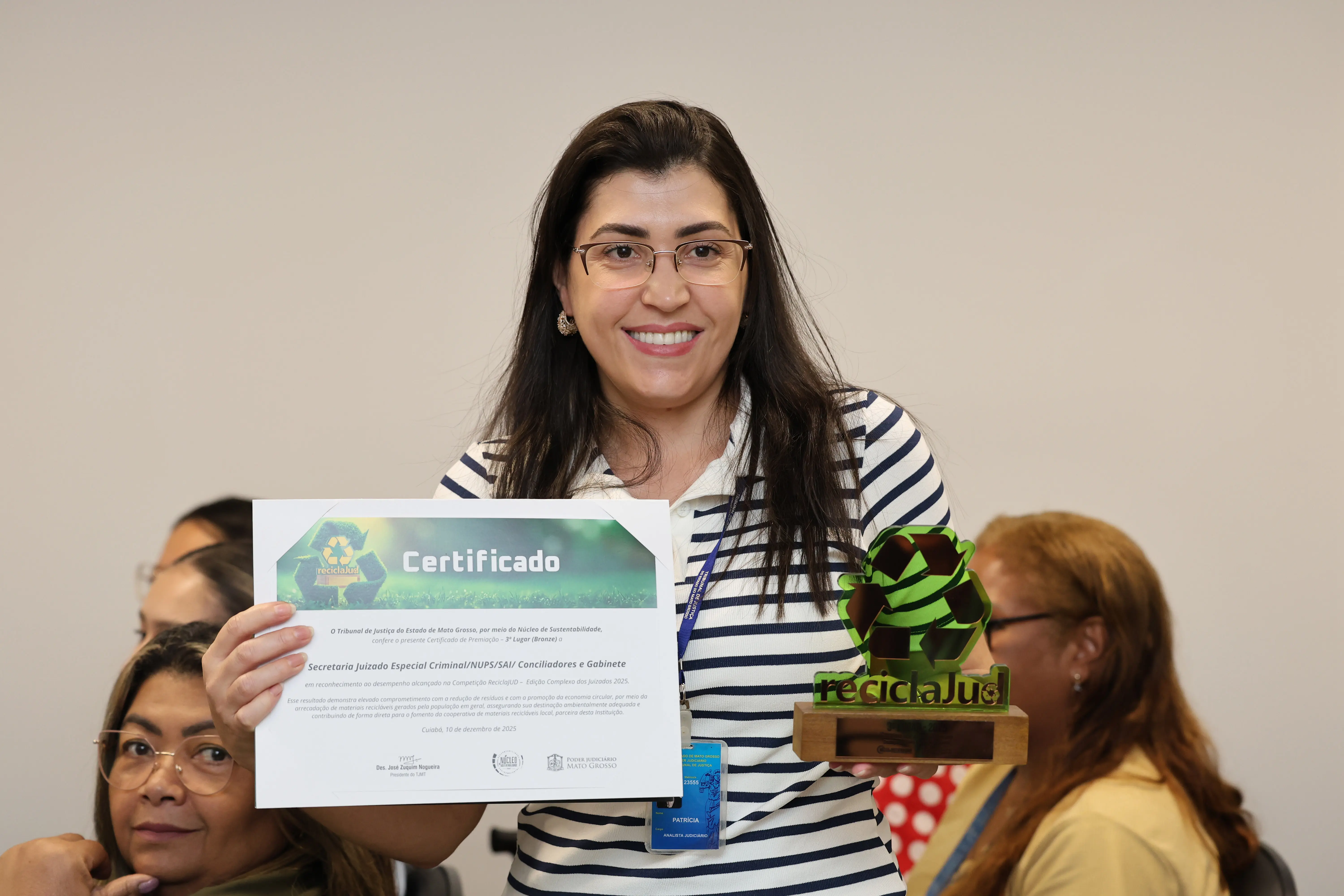 Mulher de óculos e camisa listrada segura certificado e troféu do ReciclaJud. Ela sorri diante de plateia interna. Sala do TJMT, com paredes claras e colegas sentados ao fundo. A premiação destaca desempenho sustentável.