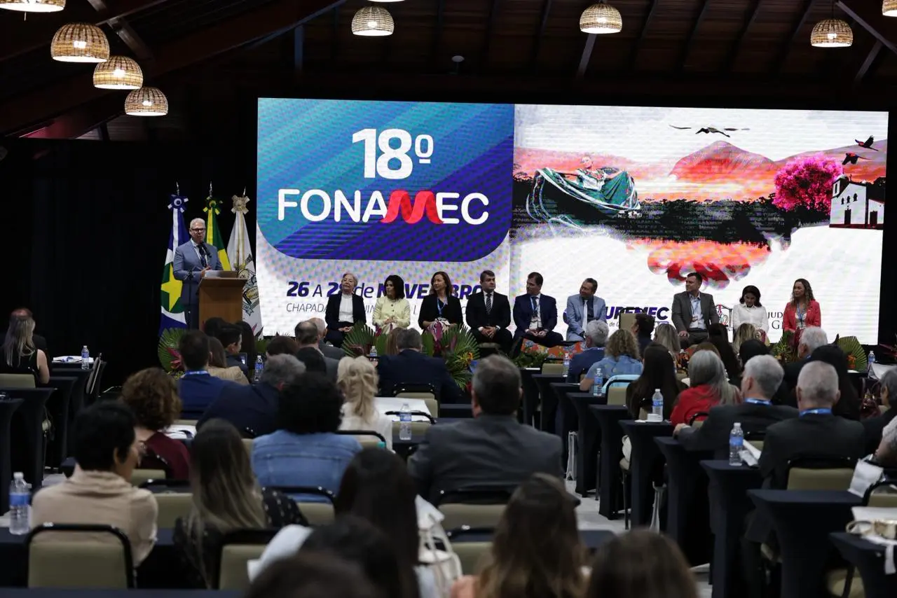 Foto em plano aberto que mostra o auditório lotado de pessoas sentadas, participando do Fonamec. No palco, várias autoridades estão sentadas e um homem fala ao microfone, no púlpito.