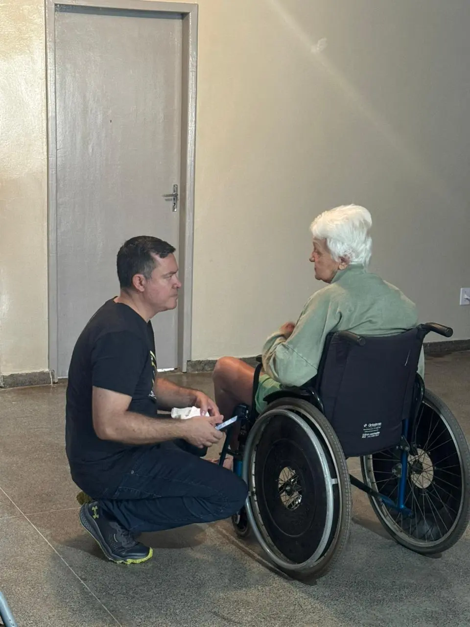 Juiz Leonísio Salles, coordenador da Justiça Comunitária se ajoelha ao lado de idosa em cadeira de rodas, conversando e oferecendo atenção. Cena ocorre em sala simples, com porta e paredes claras, reforçando acolhimento humano.