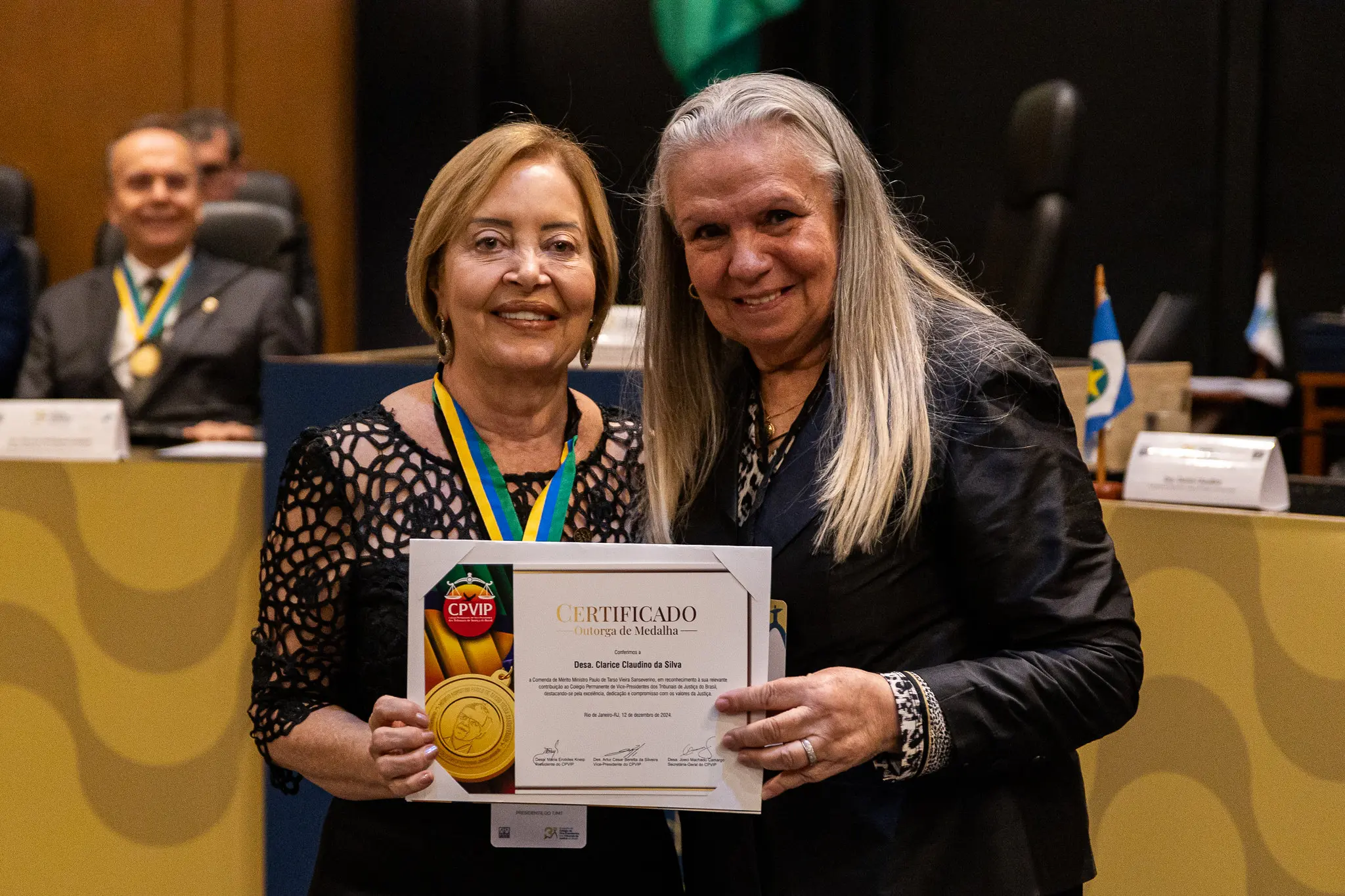 Foto em plano médio que mostra as desembargadoras Clarice Claudino e Maria Erotides Kneip posando sorrindo para a foto e segurando o certificado da Medalha do Mérito Ministro Paulo de Tarso Sanseverino, medalha que Clarice usa no pescoço.