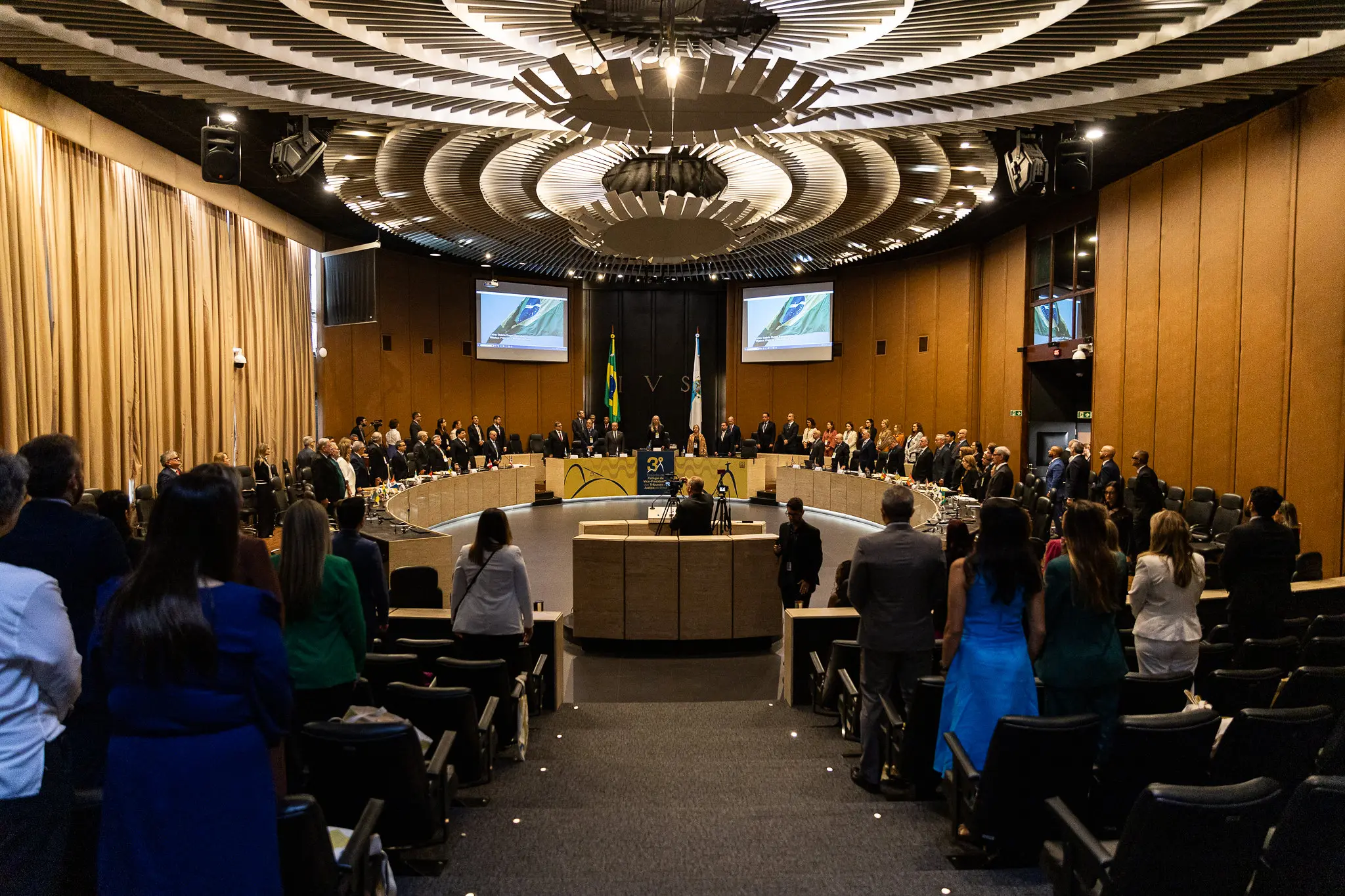Foto em plano aberto que mostra um enorme salão de conferência com uma mesa em formato de ferradura repleta de autoridades e pessoas na plateia