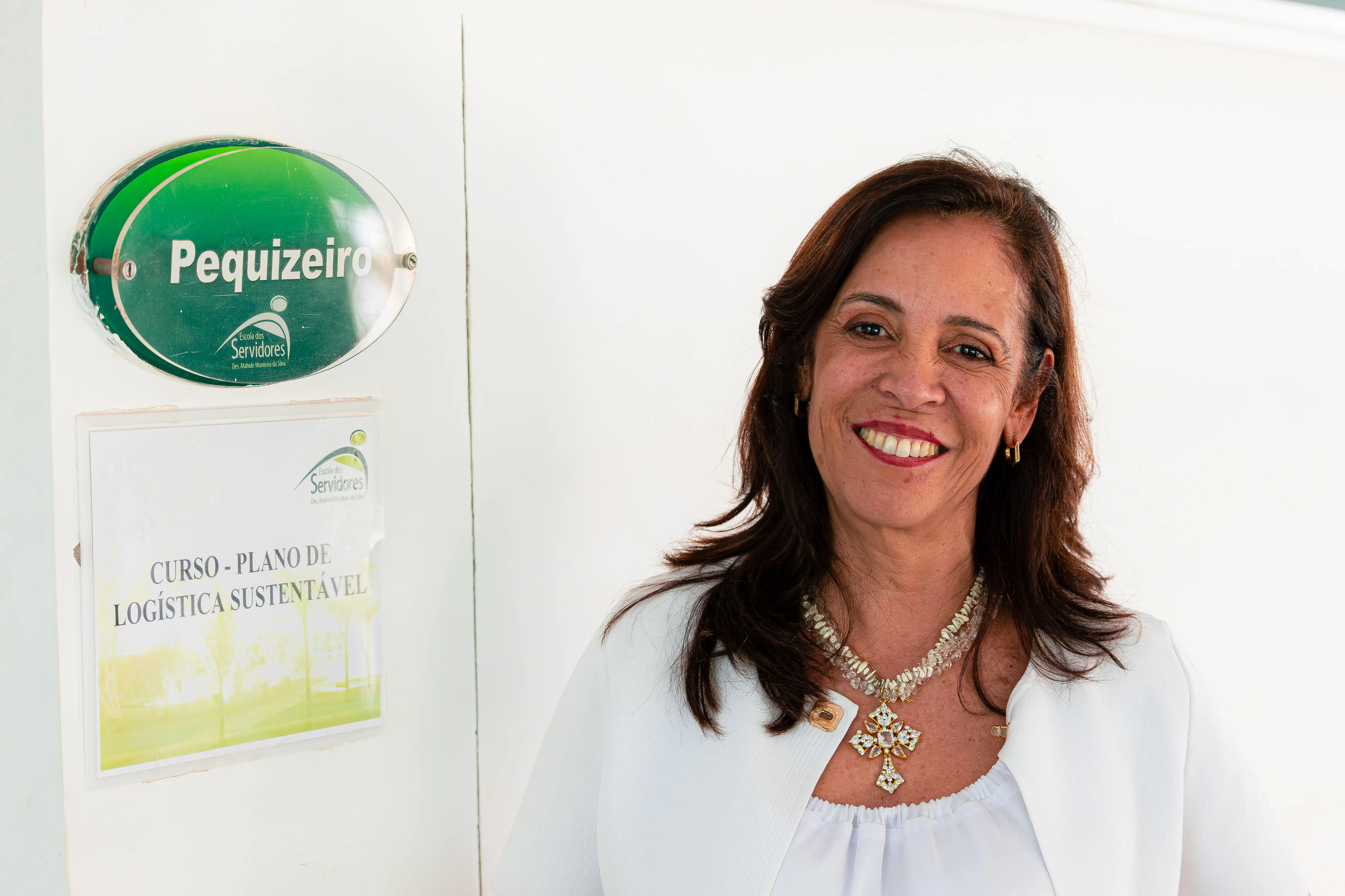 Mulher sorridente de cabelos castanhos ondulados, com blusa e casaco brancos, brincos dourados e colar de pedras com pingente grande. Está ao lado de uma parede branca com placa oval verde "Pequizeiro" e cartaz "Curso - Plano de Logística Sustentável" abaixo.