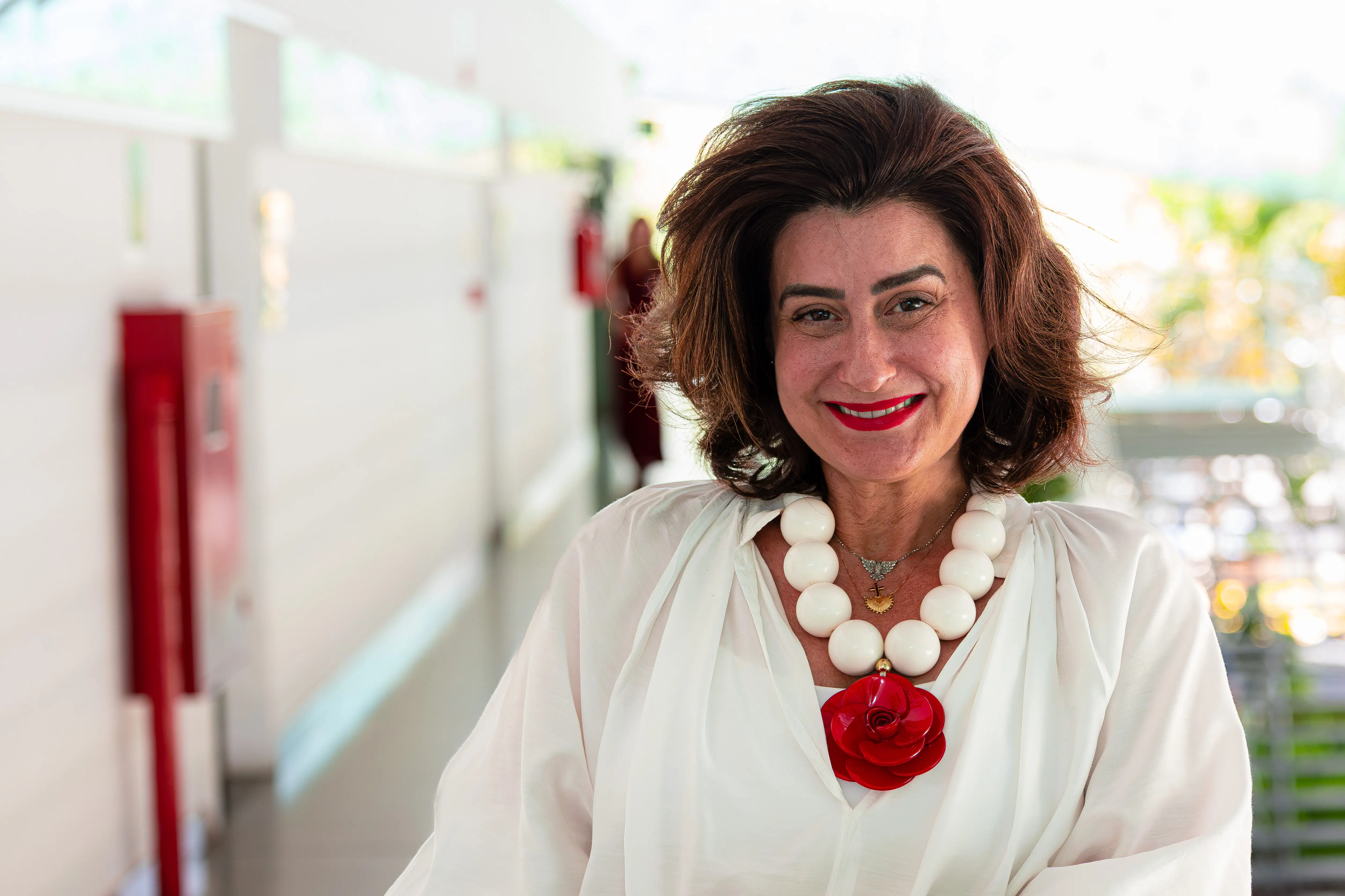 Mulher sorridente com cabelo castanho volumoso, usando blusa branca e um colar de grandes contas brancas com pingente de flor vermelha. Ela está em um corredor interno/externo, com um extintor vermelho e pessoas desfocadas ao fundo.