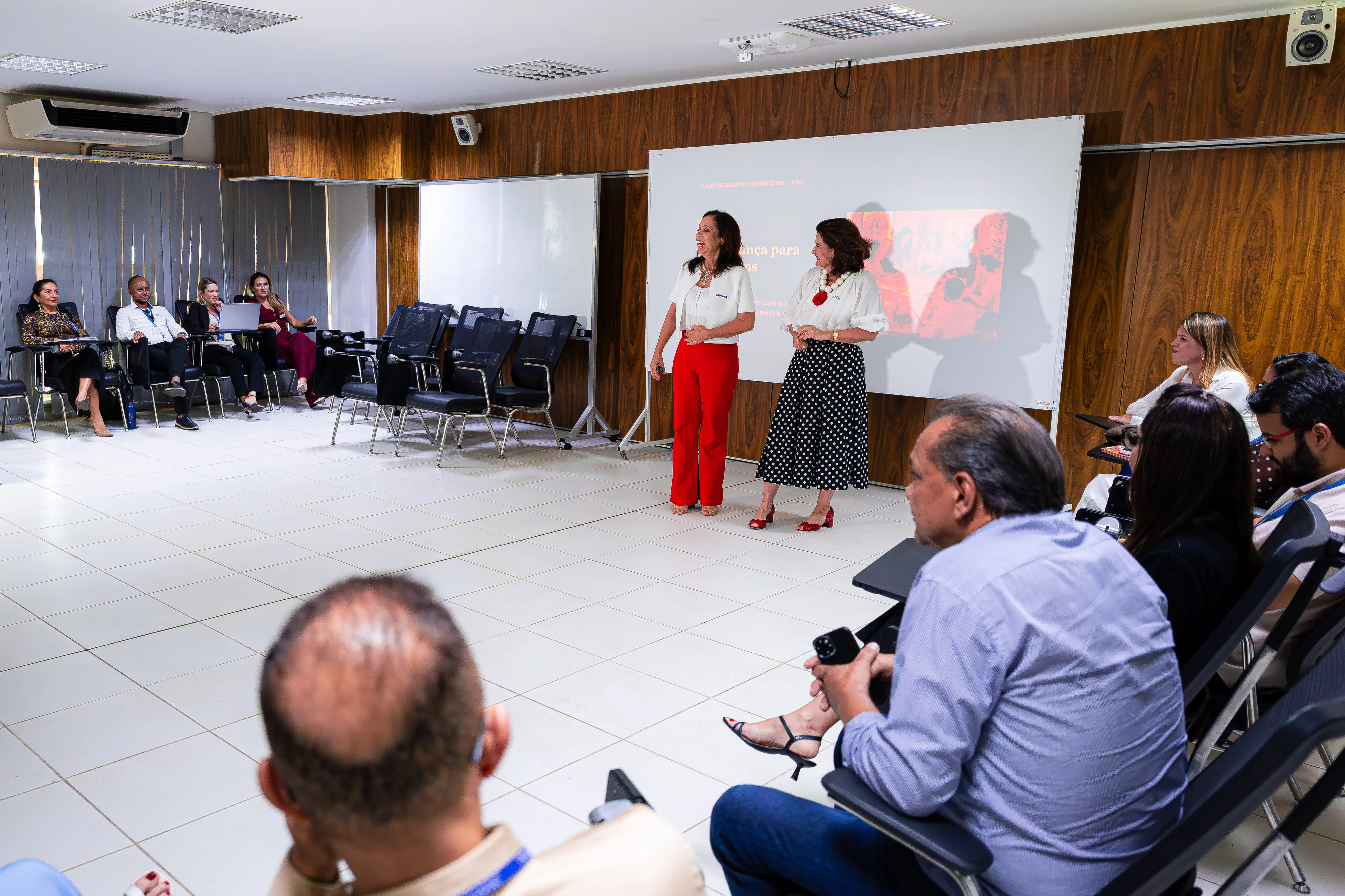 Em uma sala, duas mulheres estão de pé diante de um telão de projeção: uma veste blusa branca e calça vermelha, e a outra, blusa branca e saia de poás. À frente e à esquerda, pessoas sentadas em cadeiras as assistem. O ambiente tem piso claro e paredes com painéis de madeira.