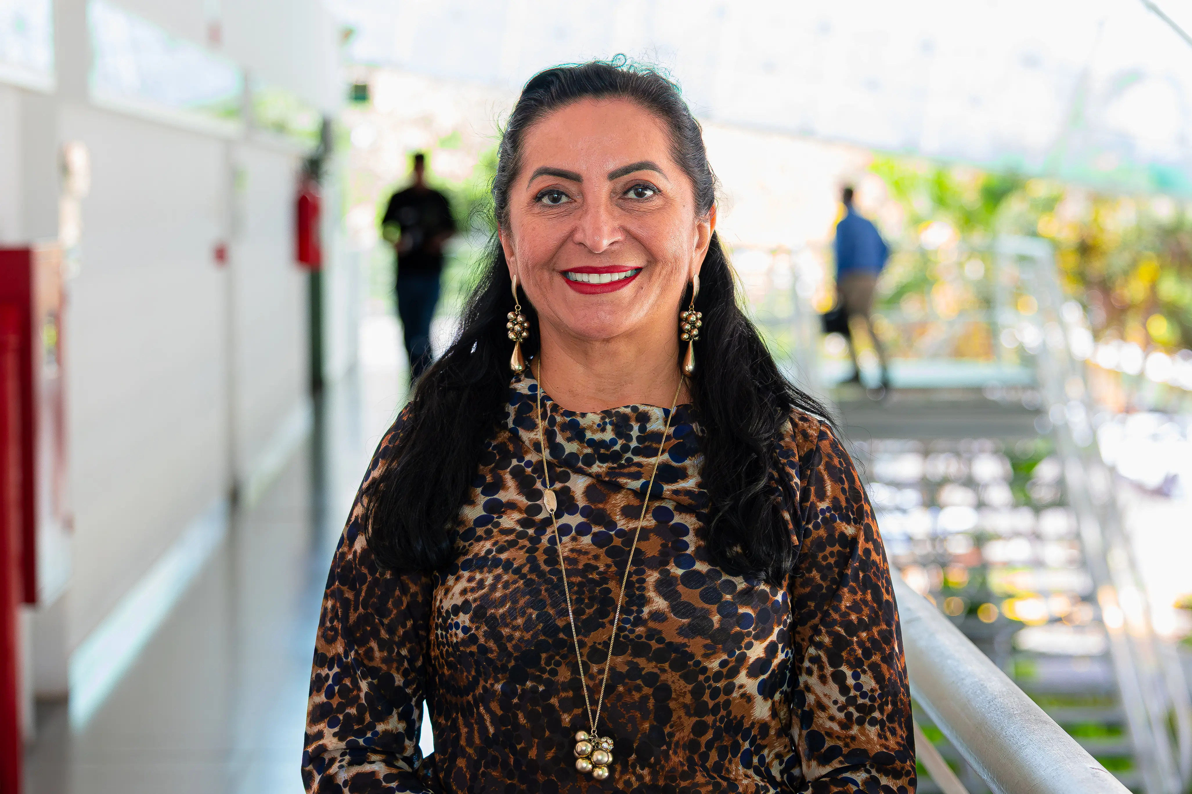 Mulher sorridente com cabelos longos escuros e ondulados, usando blusa com estampa de leopardo, brincos compridos e colar longo dourado com pingente de esferas. Ela está em um corredor ou passarela ao ar livre, com pessoas e plantas desfocadas ao fundo.