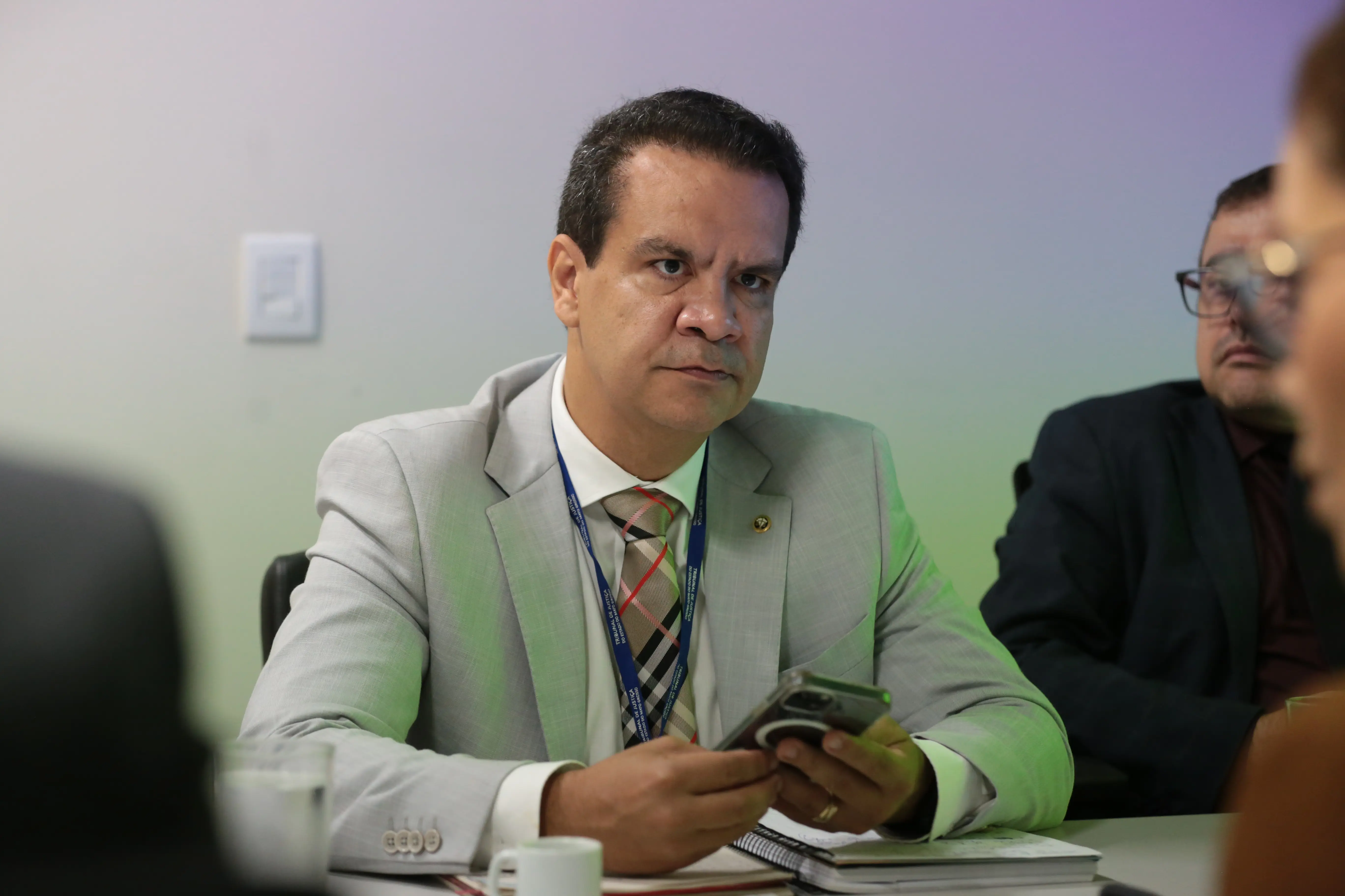 Foto que mostra o juiz Emerson Cajango sentado, participando de uma reunião. ele é um homem negro, de cabelo e olhos escuros, usando camisa branca, terno cinza e gravata xadrez nas cores marrom claro, vermelho e preto.