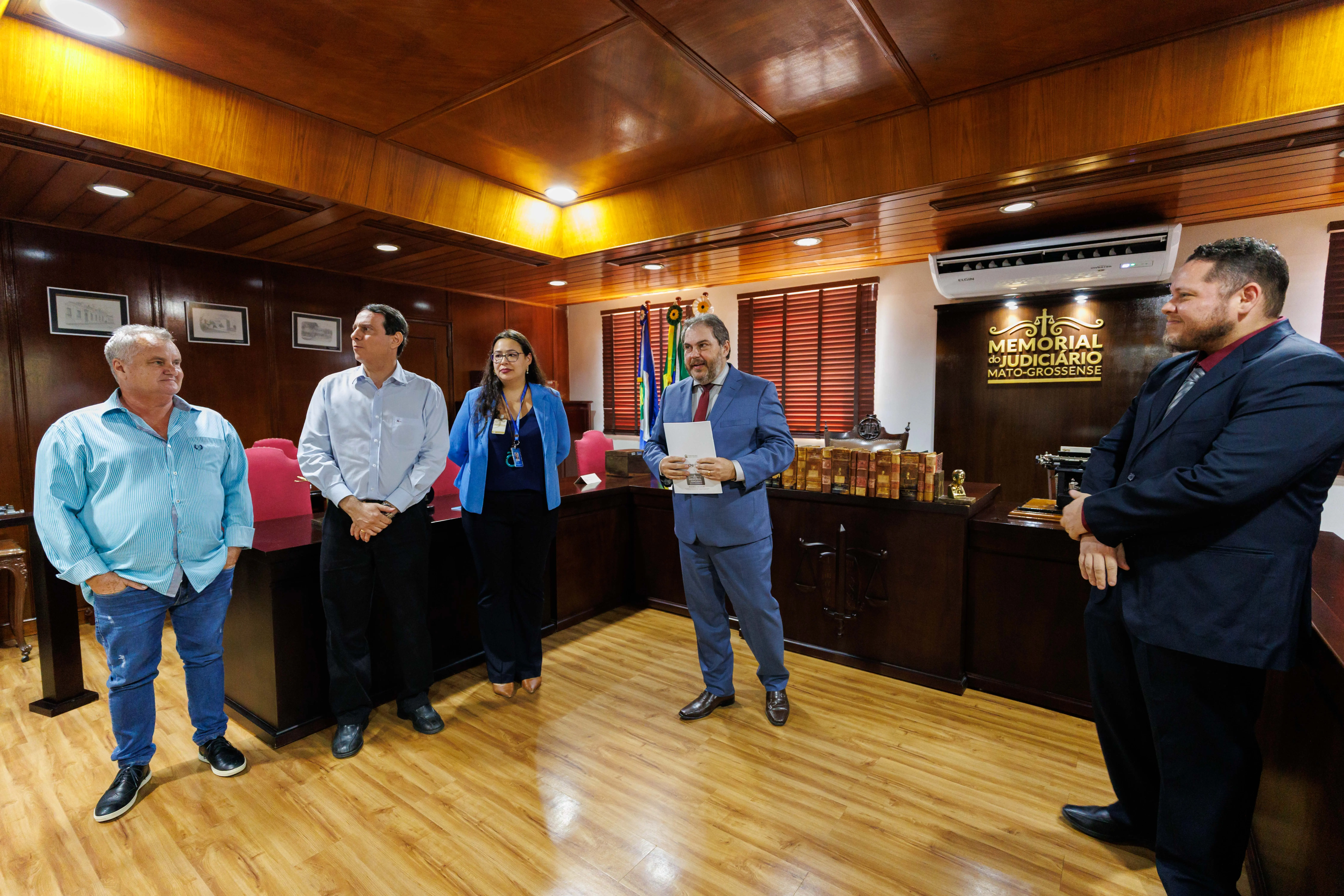 Cinco pessoas em pé no Memorial do Judiciário Mato-grossense participam de cerimônia. Ao fundo, bandeiras, livros antigos e móveis de madeira escura compõem o ambiente solene da instituição.