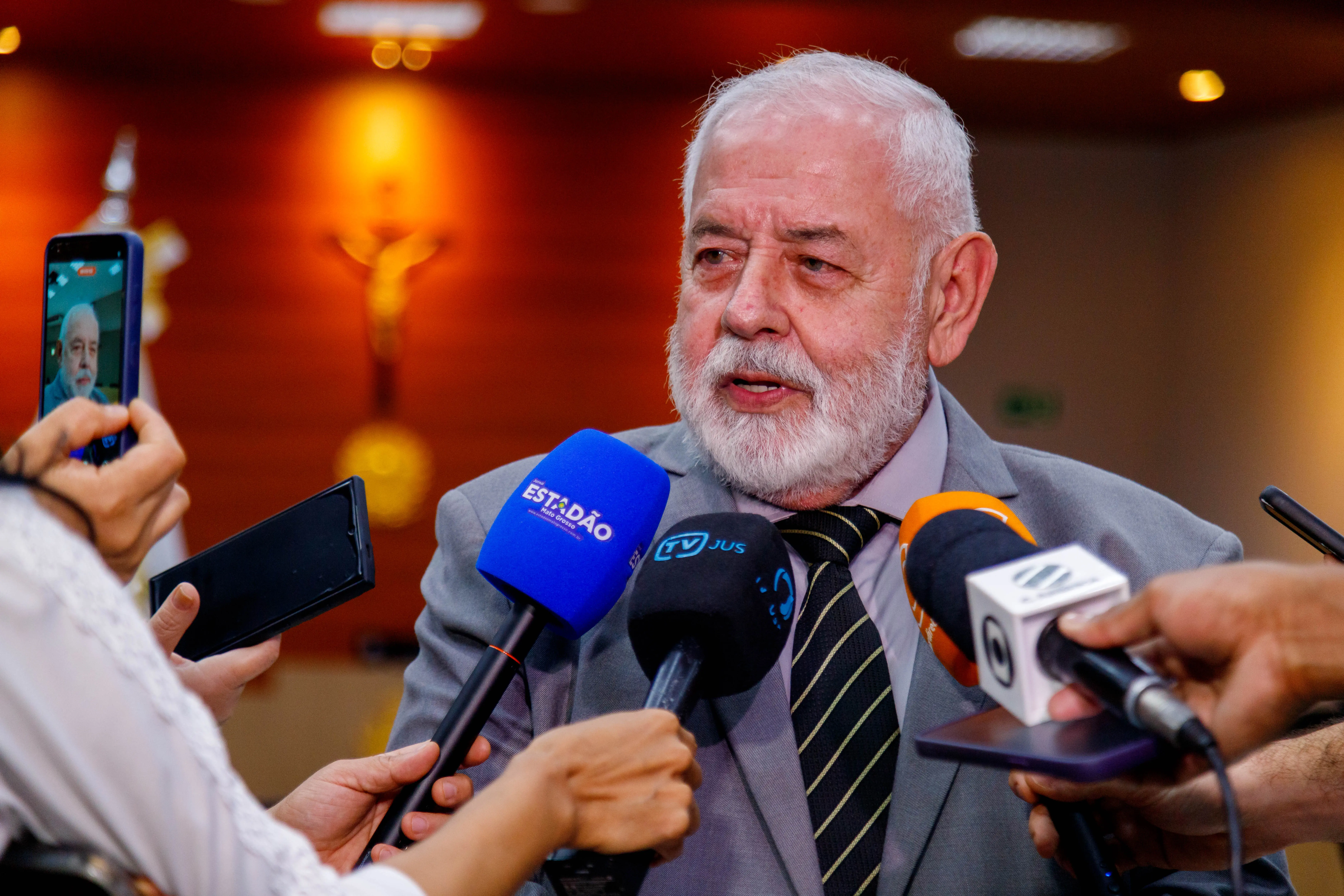 Imagem mostra um homem idoso de terno e gravata, com cabelos e barba brancos, dando entrevista. Ele está cercado por microfones de diferentes veículos de imprensa e celulares gravando. Ao fundo, há uma parede com crucifixo e iluminação suave.