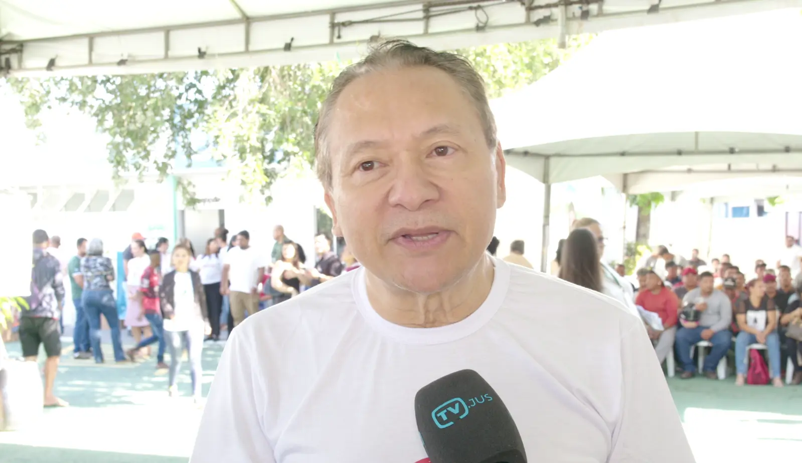 Imagem do desembargador Mário Kono durante entrevista à TV.Jus. Ele é um homem de pele clara, com traços orientais e olhos castanhos claros.  Ele veste camiseta branca do mutirão. Atrás dele é possível ver o pátio do Escritório Social com tendas e grande circulação de pessoas. 