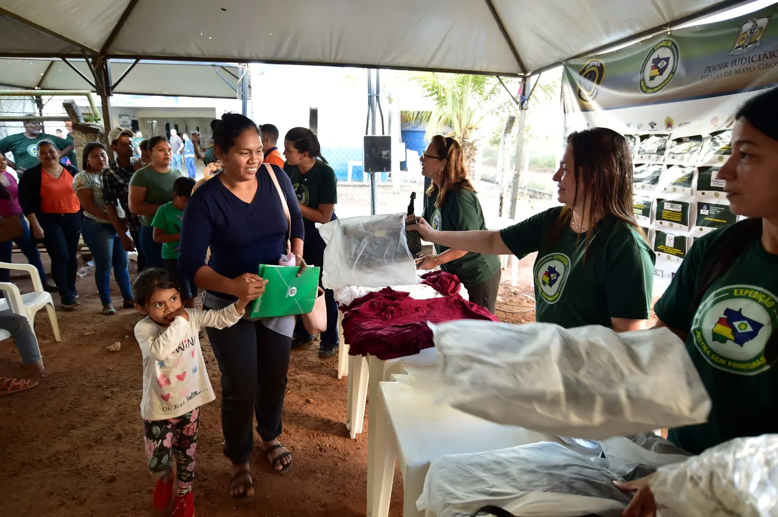 Embaixo de uma tenda, mulher segurando na mão da filha pequena recebe doação de uma bolsa das mãos de voluntária da Expedição Justiça Sem Fronteiras.