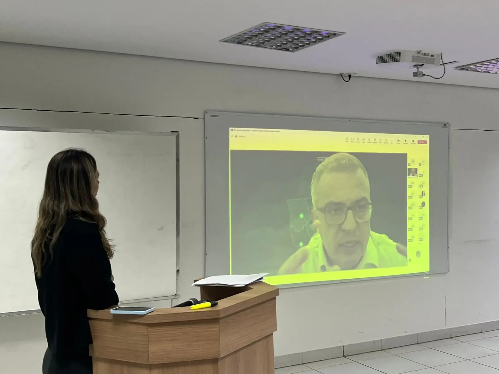 Uma mulher de costas, com cabelos longos e castanhos, está ao lado de um púlpito de madeira. Em frente a ela, uma tela exibe a imagem de um homem de óculos e camisa verde, falando.
