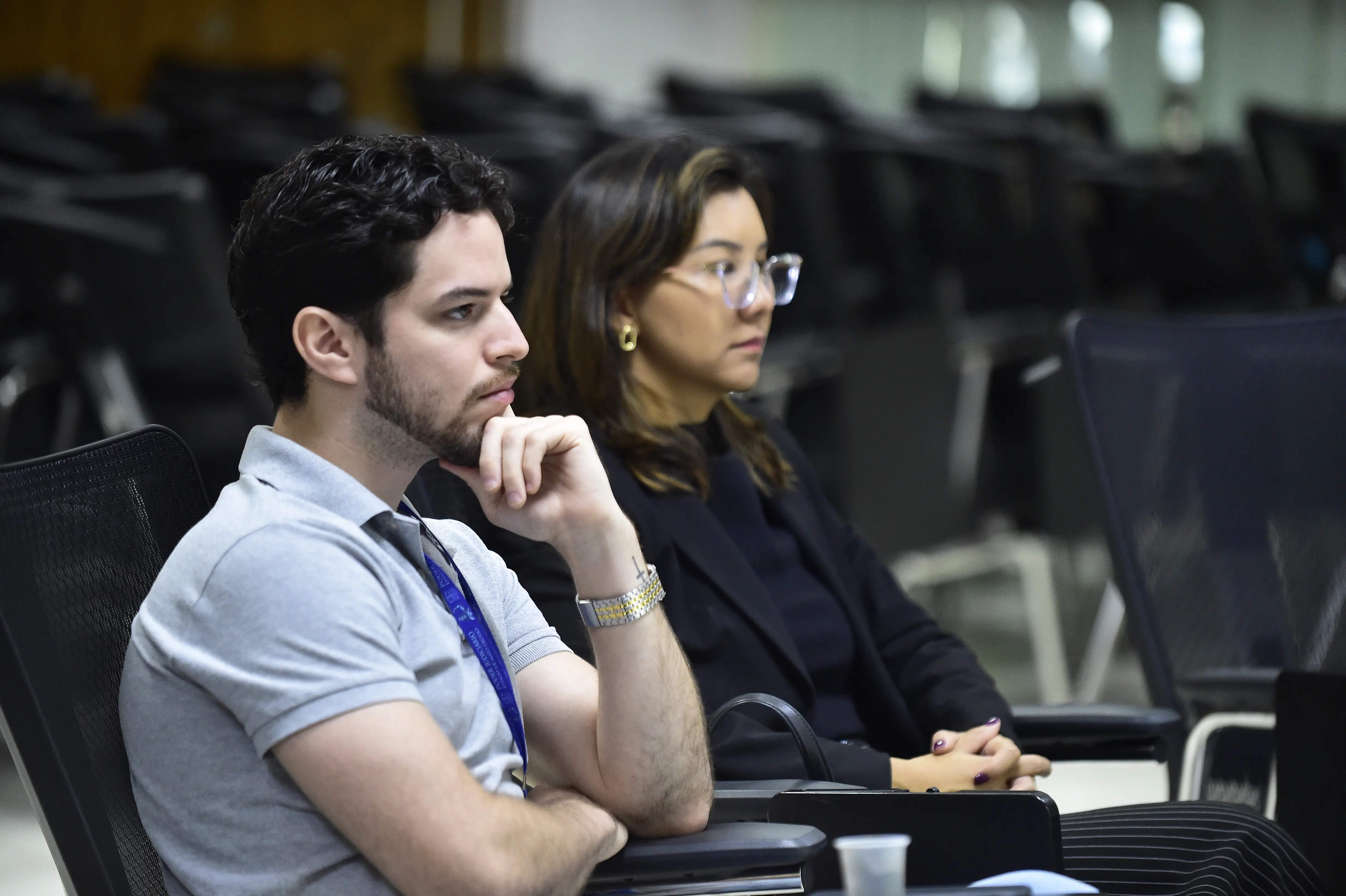 Um homem de barba e camisa polo cinza, com fone no pescoço, está sentado pensativo. Ao seu lado, uma mulher de óculos, cabelos castanhos curtos e blazer escuro o acompanha.