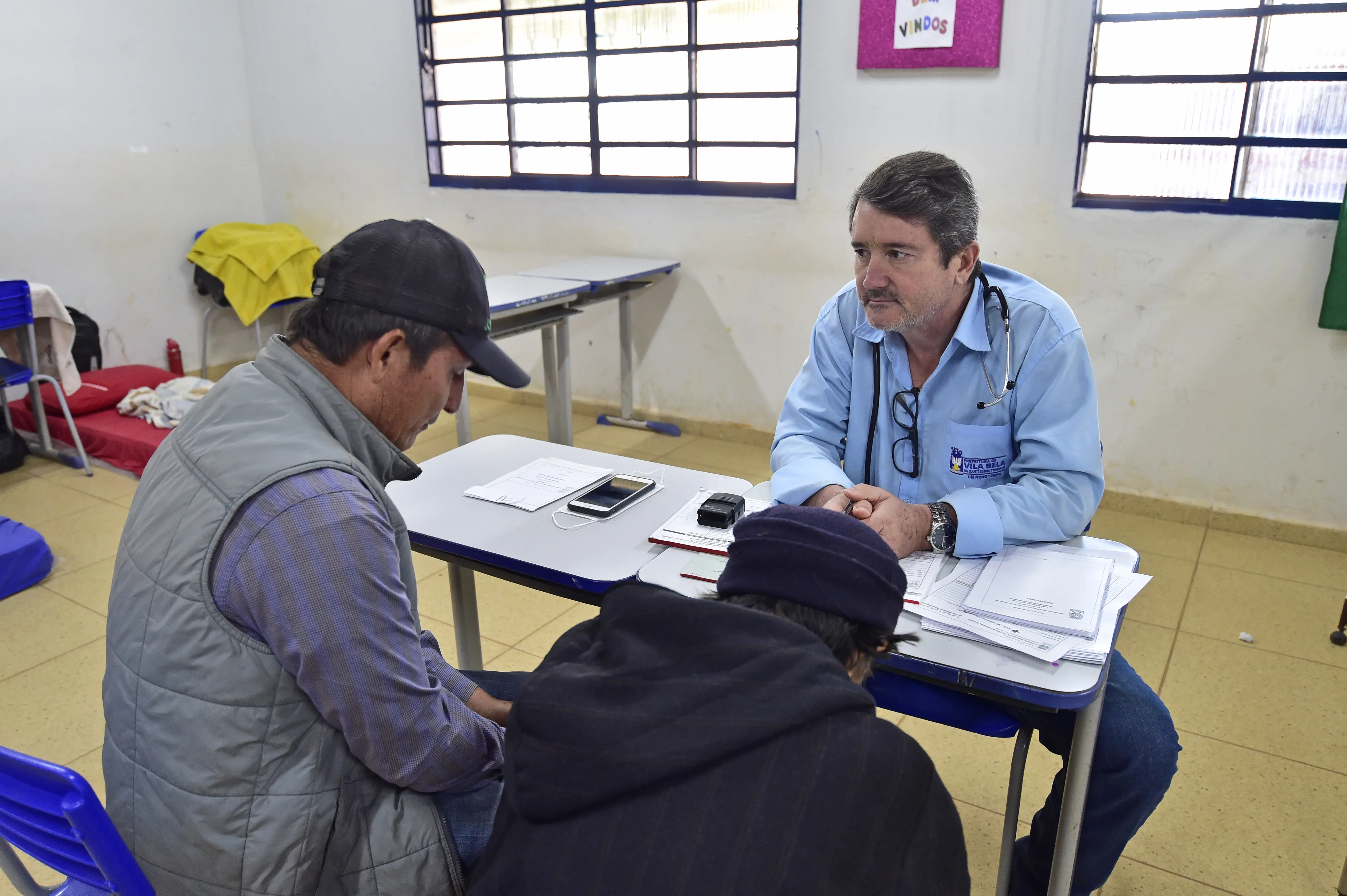 Médico usando camisa azul com logomarca da Prefeitura de Vila Bela da Santíssima Trindade atende a duas pessoas, que aparecem de costas na foto, em uma sala de aula.