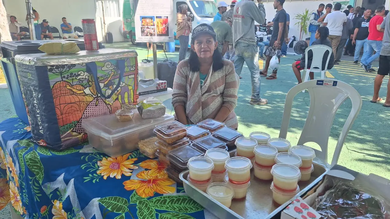 Imagem de Tânia Rocco sentada atrás de uma mesa onde seus produtos estão expostos para venda, no pátio do mutirão. Sobre a mesa, há tortas, empadas, pudins, entre outros quitutes. Tânia usa boné e blusa de frio. 