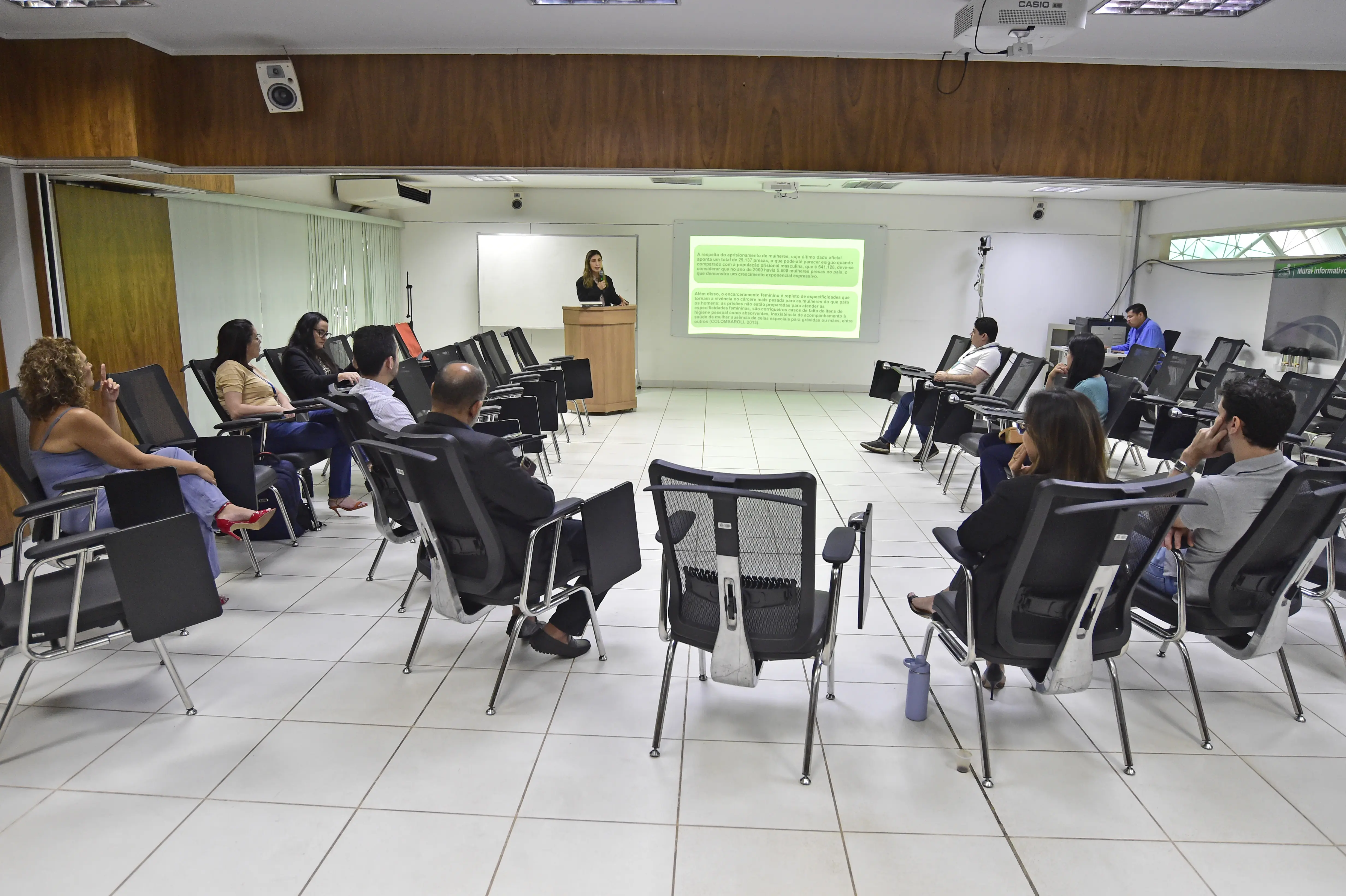 Mulher de pé discursa em auditório. Diversas pessoas, incluindo homens e mulheres, sentadas em cadeiras pretas, a assistem. Há uma projeção no fundo da sala.