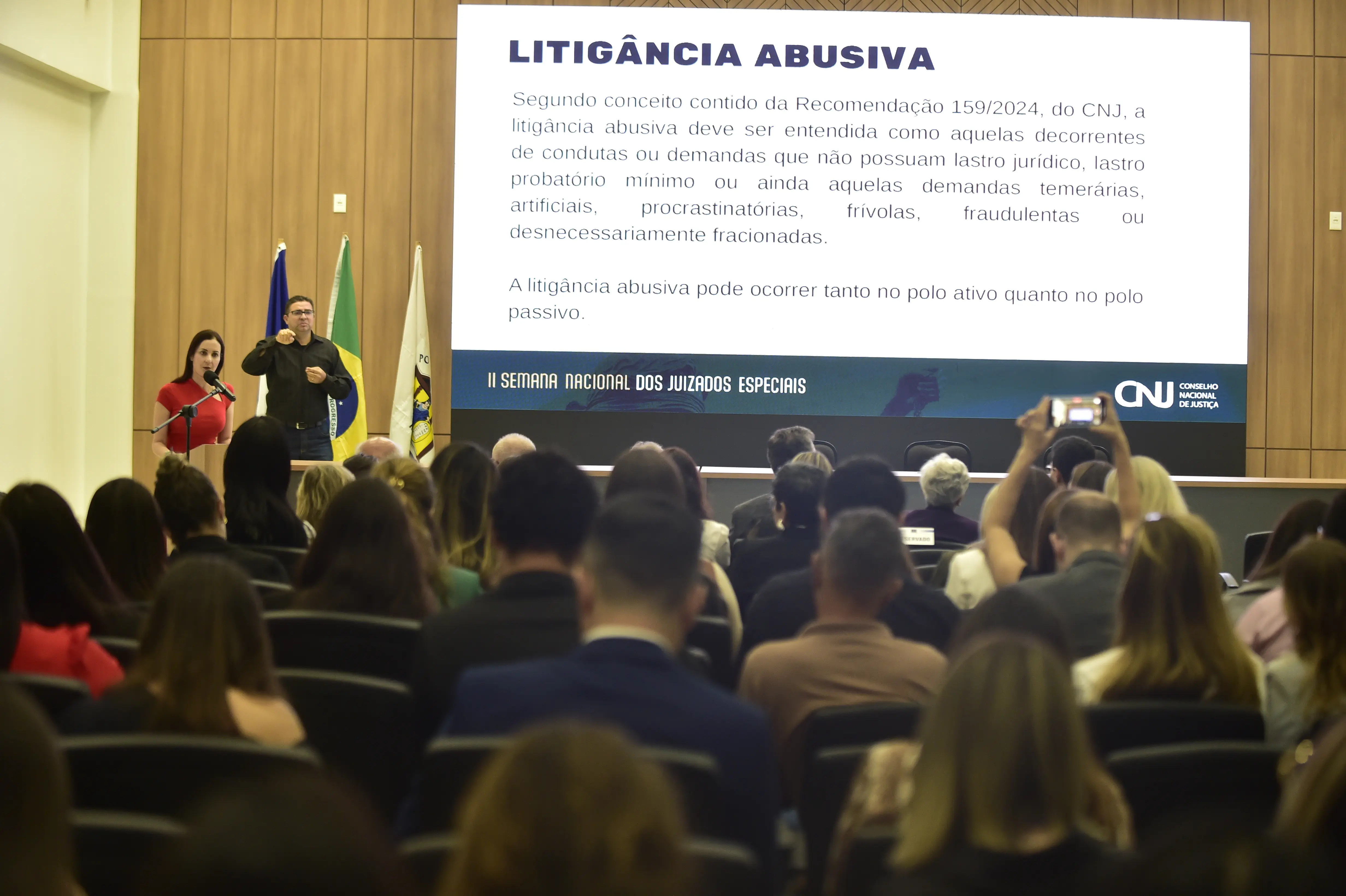 A foto mostra um auditório com pessoas sentadas, de costas, assistindo a uma apresentação. Duas pessoas estão em pé à frente, uma falando e outra gesticulando. No telão, a frase "LITIGÂNCIA ABUSIVA" em destaque.