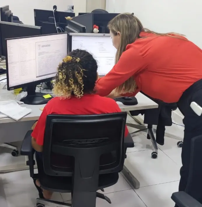 A imagem mostra uma jovem sentada em uma cadeira, trabalhando em uma mesa e recebendo auxílio de uma mulher que está ao seu lado.