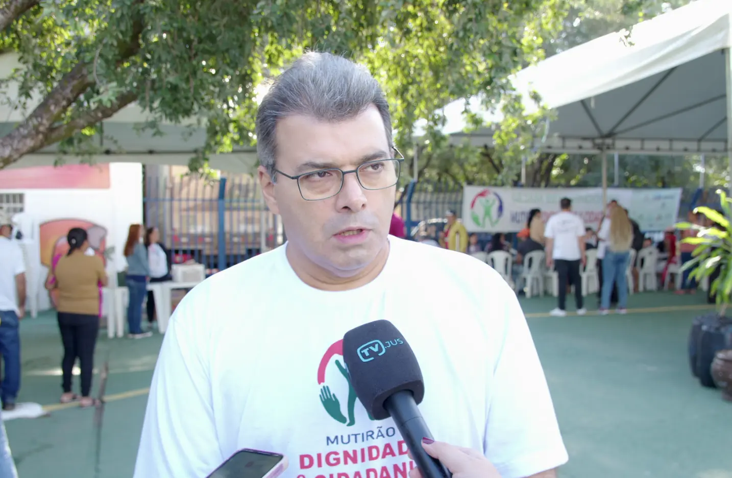 Imagem do juiz Geraldo Fidelis durante entrevista á TV.Jus. Ele é um homem branco, de cabelos grisalhos, usando óculos de grau e camiseta branca com estampa do mutirão. Atrás dele, é possível ver um pátio com tendas e pessoas circulando. 