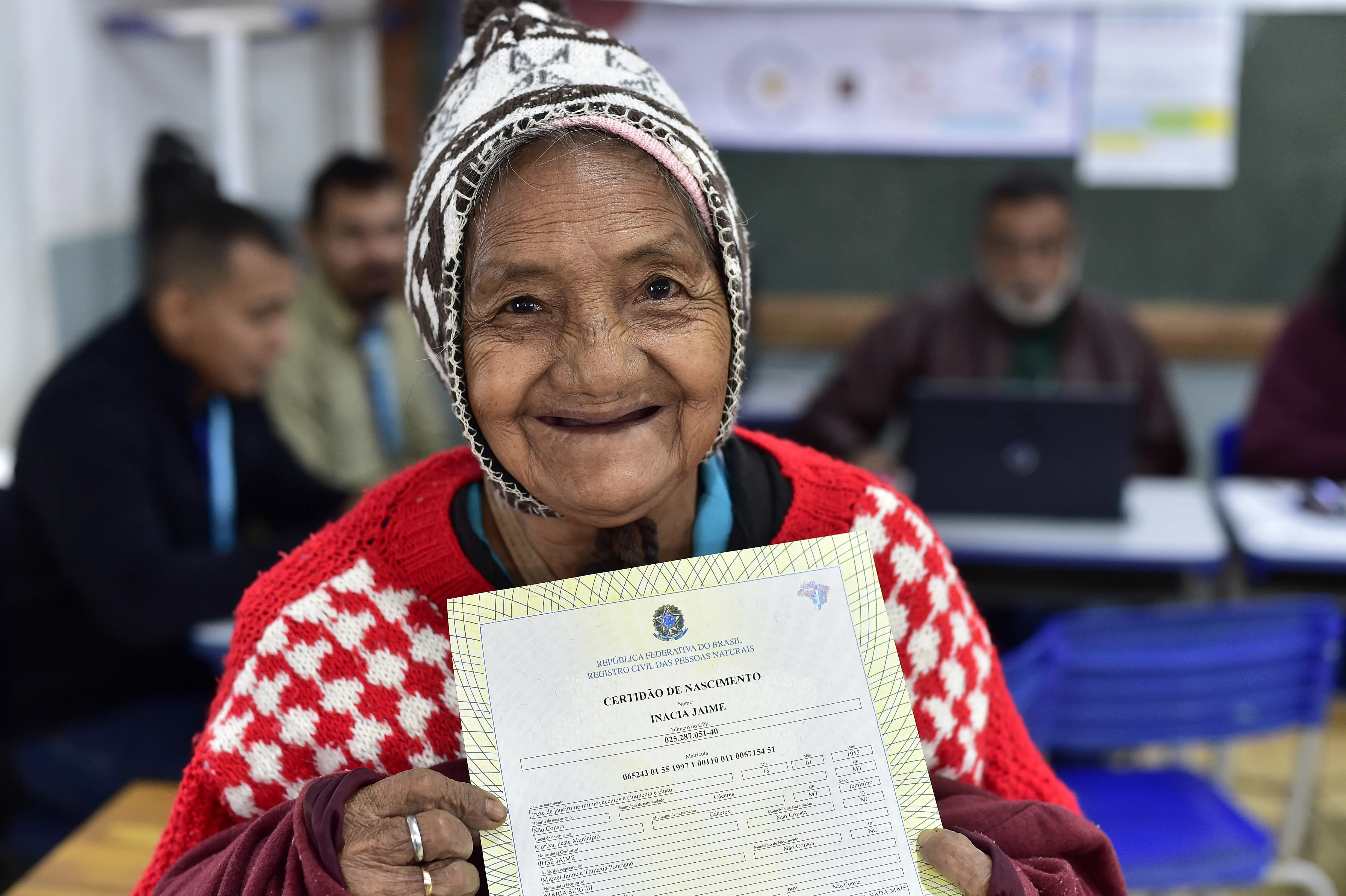 Uma pessoa segura uma Certidão de Nascimento com as duas mãos. Ela veste um gorro com padrões coloridos e um suéter vermelho com detalhes brancos. Ao fundo, há várias pessoas sentadas em mesas, algumas utilizando laptops. Os rostos estão desfocados