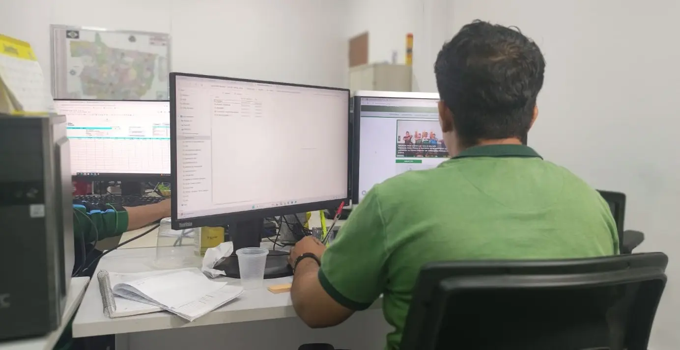 A imagem mostra uma pessoa de costas, sentada em uma cadeira e trabalhando em uma mesa com dois monitores.