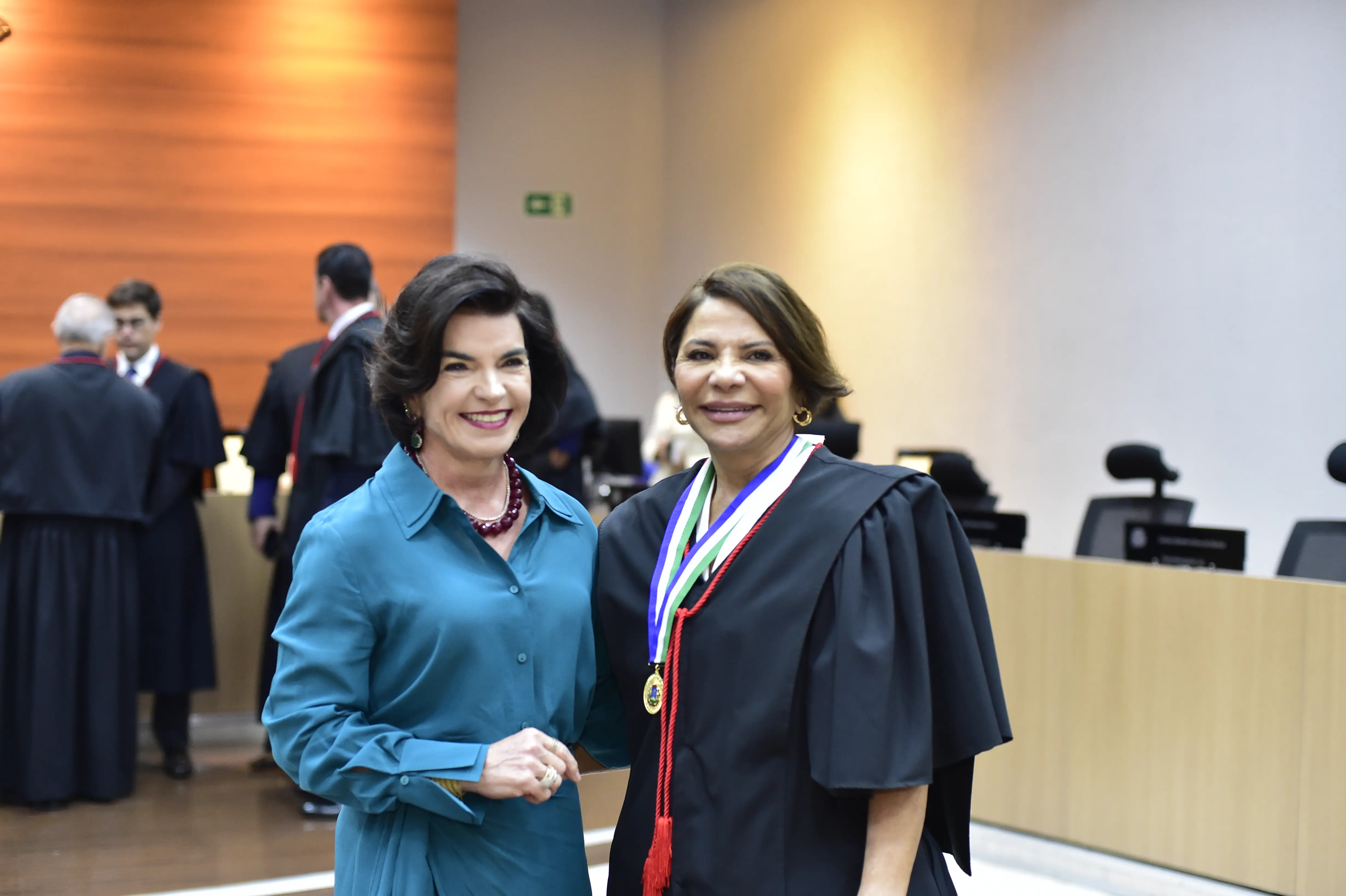 Duas mulheres adultas sorriem para a câmera em um ambiente interno e formal. A da esquerda veste uma camisa azul-petróleo e tem cabelo escuro curto. A da direita usa uma toga preta e uma medalha colorida no pescoço. O fundo é desfocado, com pessoas e móveis de escritório.