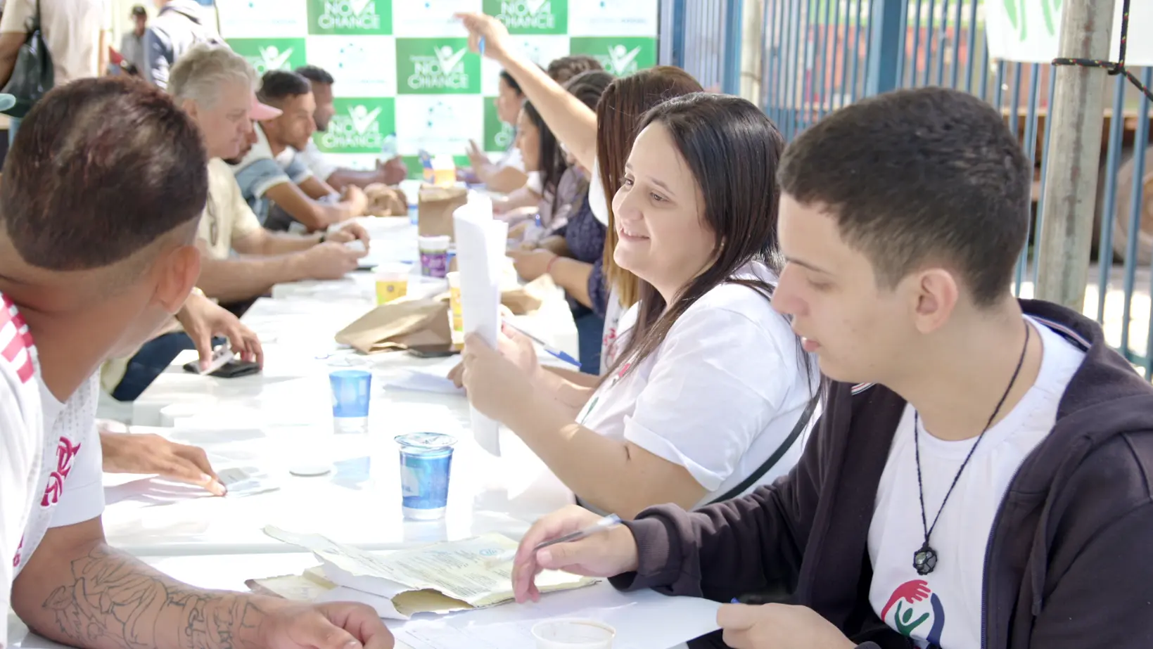 Imagem que mostra uma mesa grande com várias pessoas sendo atendidas no Mutirão Dignidade e Cidadania. Em primeiro plano aparece uma moça do lado direito da mesa olhando para o rapaz do outro lado da mesa e sorrindo. Ela segura papeis e uma caneta. 
