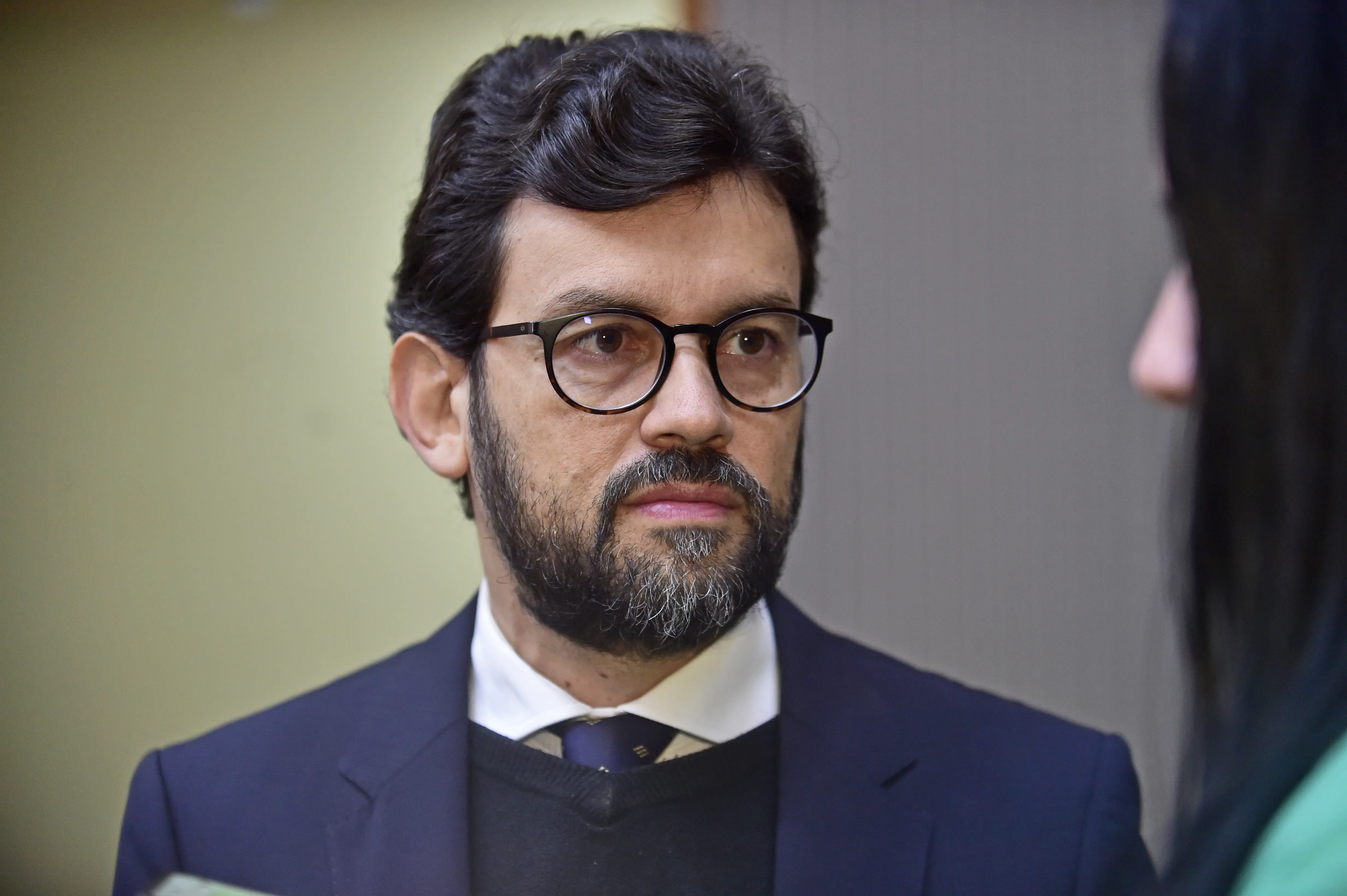 Juiz Tiago Souza Nogueira de Abreu tem pele clara, com cabelos escuros e barba. Ele usa óculos de armação redonda, um terno azul-escuro sobre um suéter preto e uma camisa branca com gravata estampada