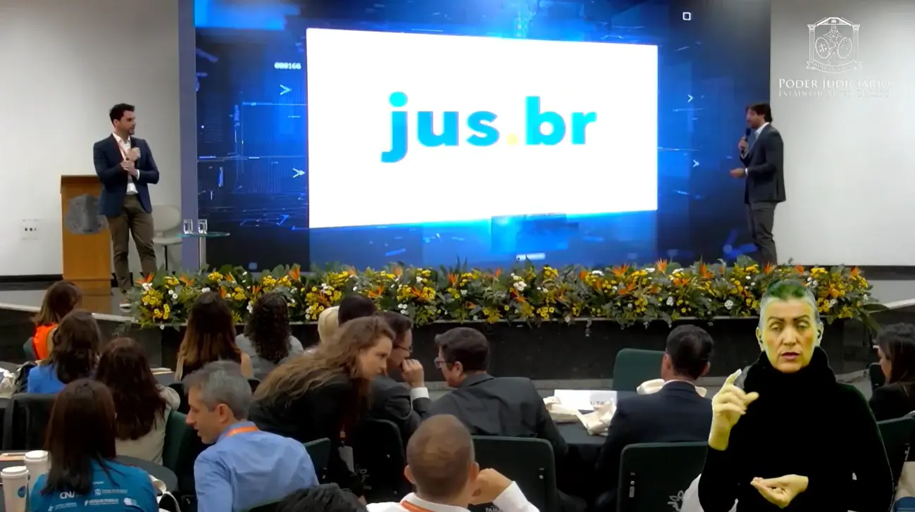 Print da transmissão da apresentação do Jus.br. A imagem mostra dois juízes no palco do evento. Ao centro, um grande telão com a logomarca do Jus.br. Na parte inferior da imagem, dezenas de participantes assistem à apresentação, sentados em mesas espalhadas no salão. 