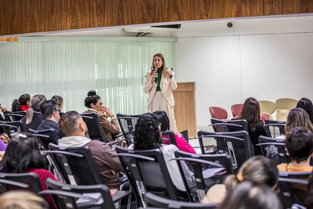 Mulher loira em palco discursa com microfone, vestindo blazer creme e blusa verde. Audiência atenta em cadeiras escuras, com costas visíveis. O ambiente possui paredes de madeira e painel branco com cortinas e luminárias.