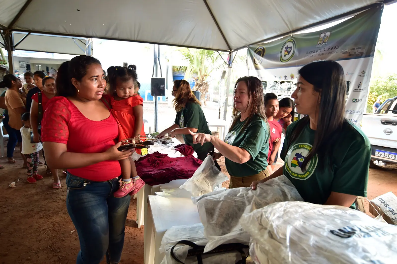 Embaixo de uma tenda, mulher com menina no colo recebe doação de calçado das mãos de voluntária da Expedição Justiça Sem Fronteiras.