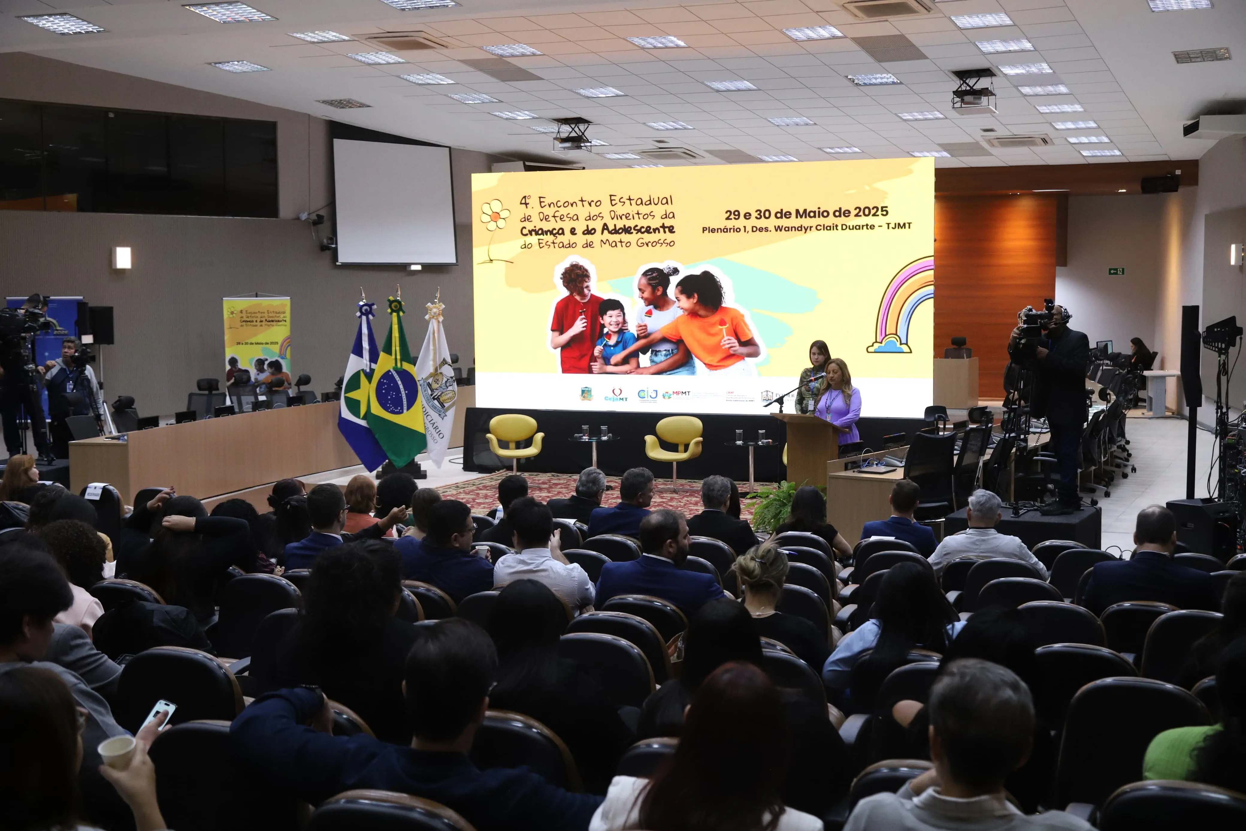 A imagem mostra um auditório amplo e escuro, repleto de pessoas sentadas de costas, assistindo a um grande palco iluminado. No palco, um telão vibrante exibe "4º Encontro Estadual de Defesa dos Direitos da Criança e do Adolescente do Estado de Mato Grosso", com a data "29 e 30 de Maio de 2025".