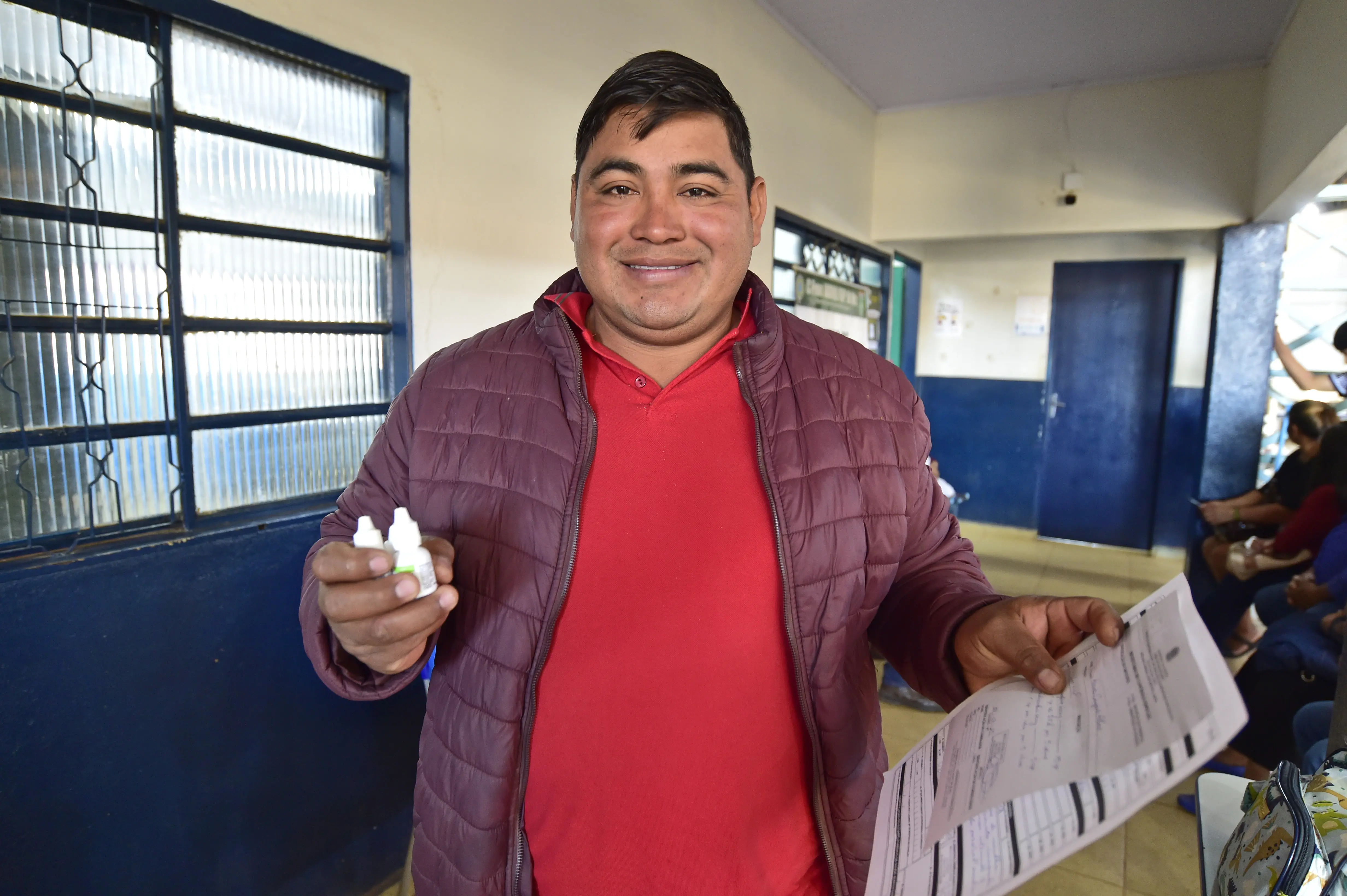 Homem sorrindo mostra dois frascos de remédios e, na outra mão, receita médica. Ele é negro, de cabelos lisos e pretos, com traços bolivianos, usando camiseta vermelha e jaqueta vinho. ele está no pátio de uma escola.