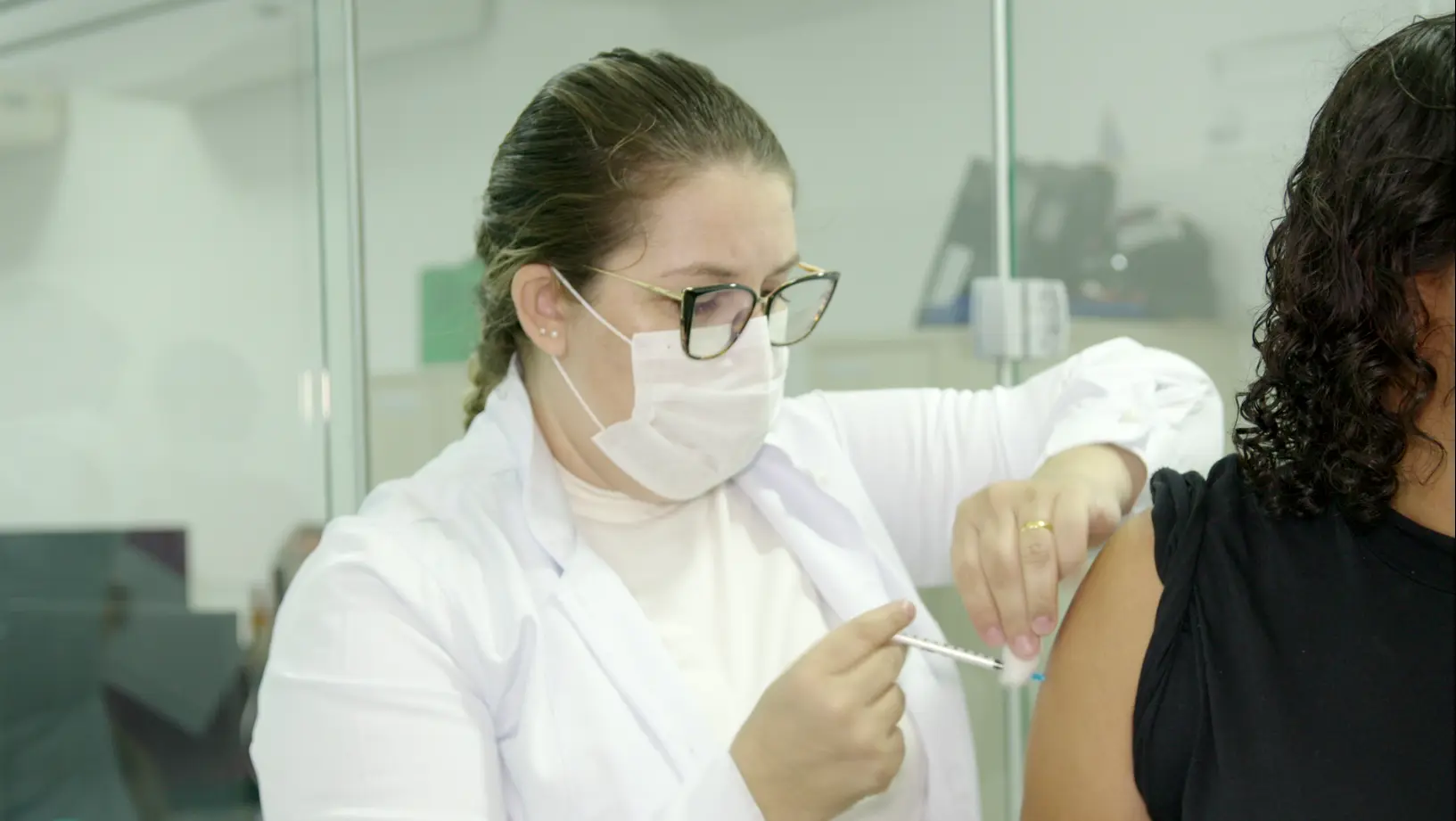 Imagem de uma enfermeira aplicando vacina no braço de uma mulher. A enfermeira é branca, de cabelos loiros e com trança, usando jaleco branco e máscara. A mulher que recebe a vacina tem cabelos escuros e cacheados e usa blusa preta. 