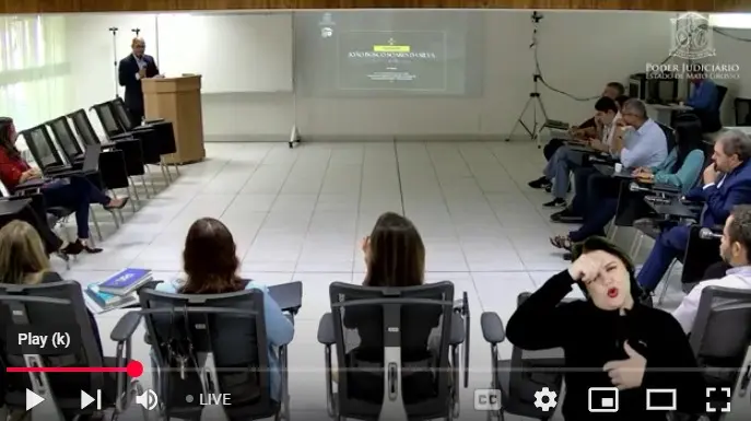 print de tela da sala de aula, onde os alunos estão sentados em cadeiras dispostas em U e o professor fala ao púlpito. 