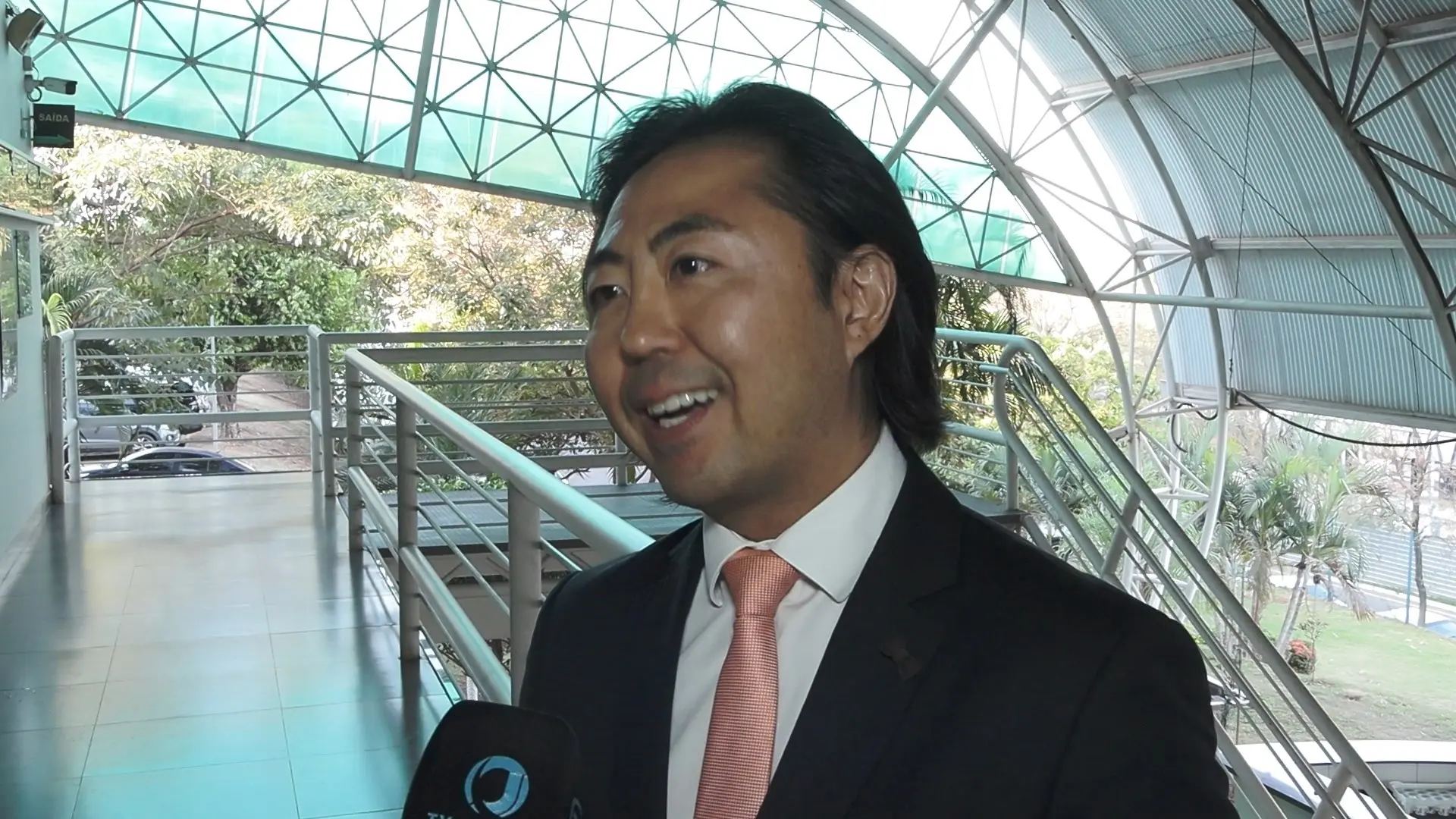 juiz Fernando Kendi Ishikawa usa terno escuro e gravata salmão fala sorridente em entrevista para TV Justiça. Ele está em área envidraçada e coberta, com árvores ao fundo, transmitindo leveza e formalidade.