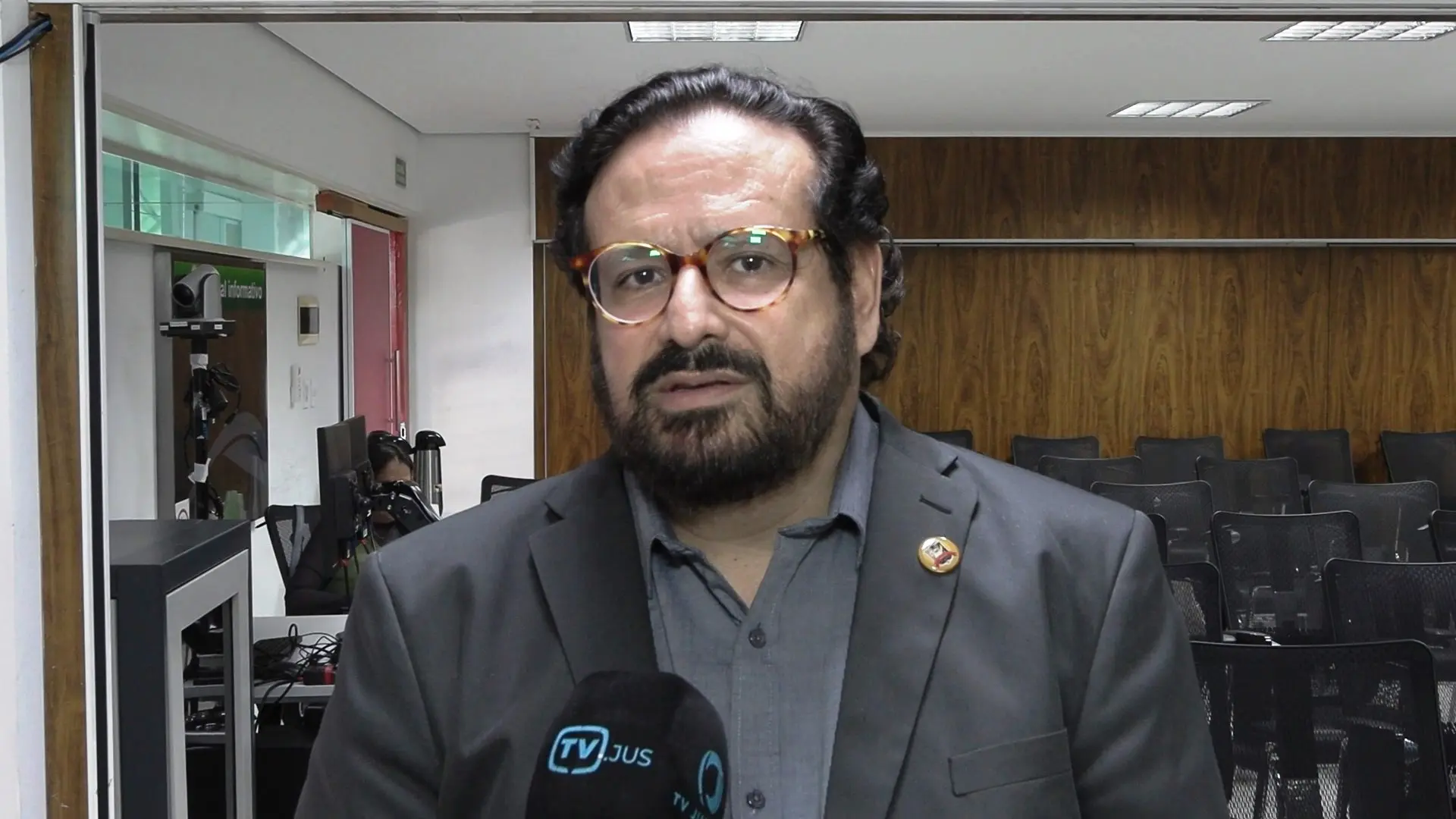 juiz Gonçalo Antunes de Barros usa óculos e barba escura dá entrevista para TV Justiça. Usa terno preto e camisa cinza, com crachá preso ao paletó. Ao fundo, auditório com cadeiras vazias e painel de madeira.