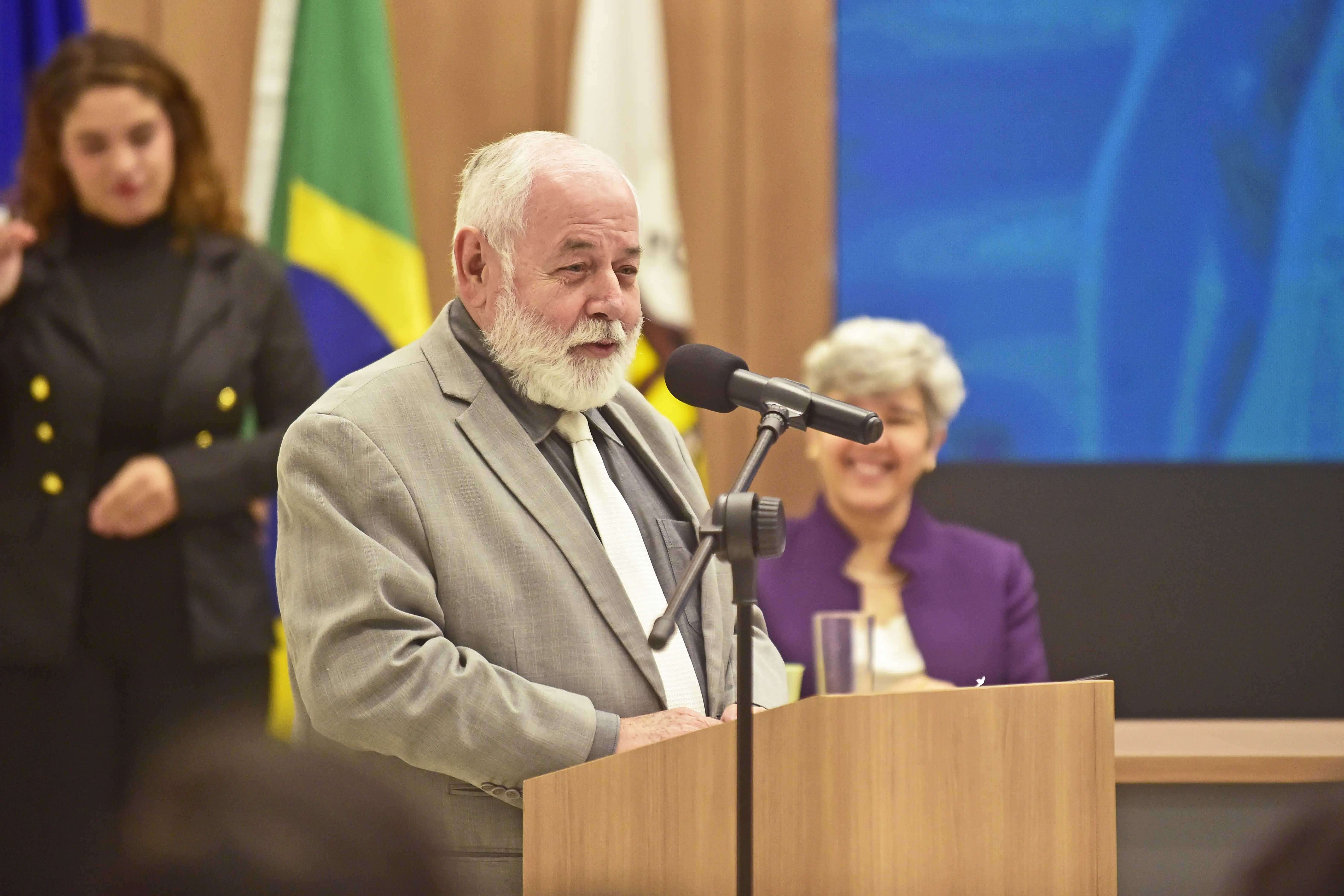 Desembargador José Zuquim é um homem branco, com cabelos e barba brancos, está de pé em um púlpito de madeira, falando em um microfone. Ele usa um terno cinza e uma gravata branca. 