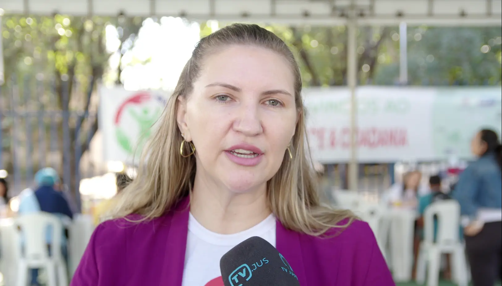 Imagem de Beatriz Dziobat concedendo entrevista à TV.Jus. Ela é uma mulher branca e loira, usando camiseta do mutirão e blazer roxo. 