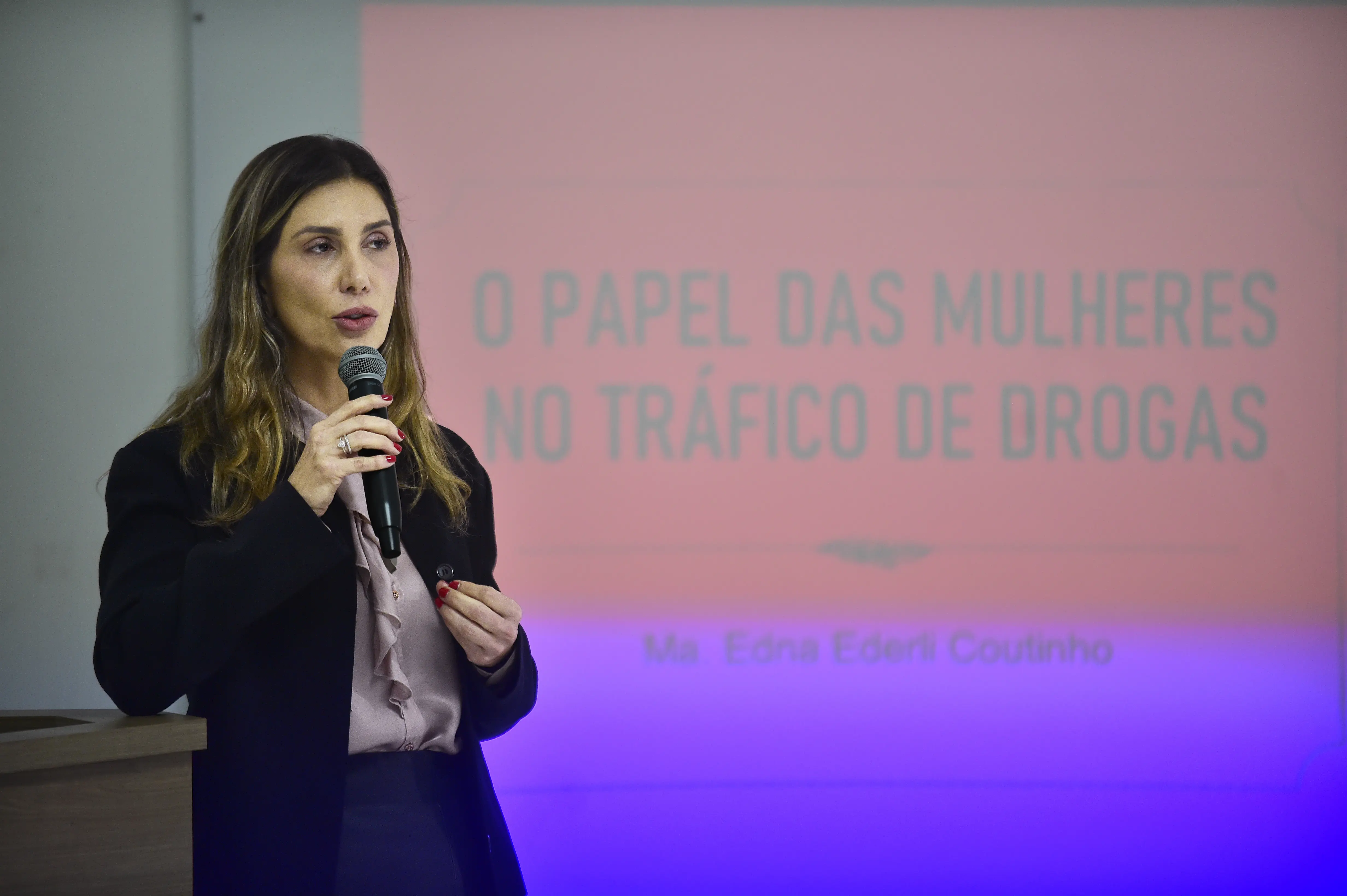 Uma mulher com cabelos castanhos longos e blusa rosa, fala ao microfone. Atrás dela, um telão projeta: "O PAPEL DAS MULHERES NO TRÁFICO DE DROGAS - Ma. Edna Ederli Coutinho".