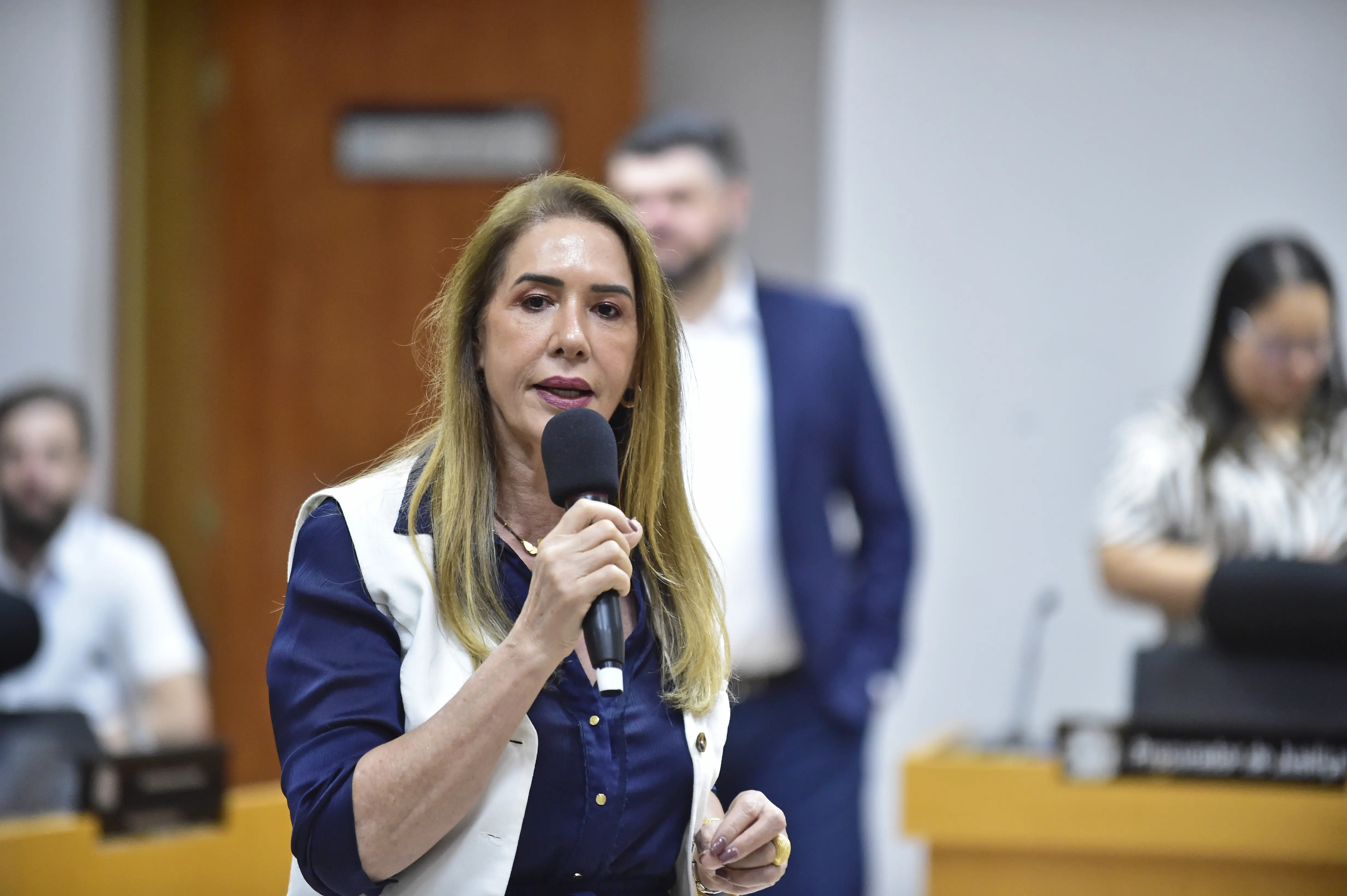 Mulher segura microfone e fala durante evento institucional. Ela veste camisa azul com colete branco. Ao fundo, pessoas acompanham a atividade em ambiente formal, com mesas, placas e microfones.