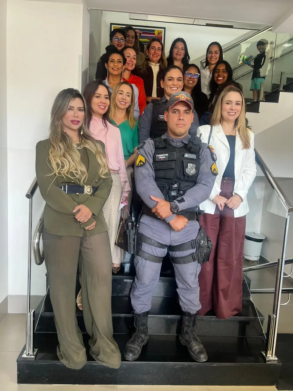 Foto na vertical que mostra vários integrantes da Rede de Enfrentamento à Violência Doméstica de Cuiabá (a maioria mulheres) posando e sorrindo para a foto. Eles estão em pé em uma escada. À frente do grupo de pessoas, estão a delegada Judá Marcondes, um policial militar e a juíza Ana Graziela Vaz.