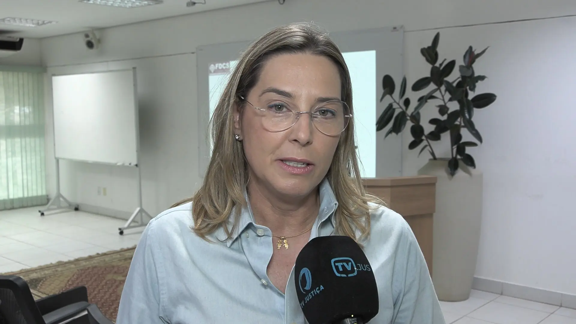juíza Gabriela Knaul é loira, de óculos, fala em entrevista à TV Justiça. Usa camisa azul clara e colar discreto. Atrás, painel de apresentação sobre independência judicial e vaso decorativo próximo ao púlpito.