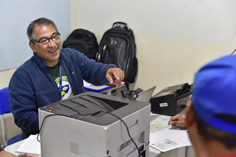 Servidor da Receita Federal atende a um homem, sorrindo. Ele é um senhor usando óculos de grau e moletom azul escuro por cima da camiseta verde da Justiça Sem Fronteiras.