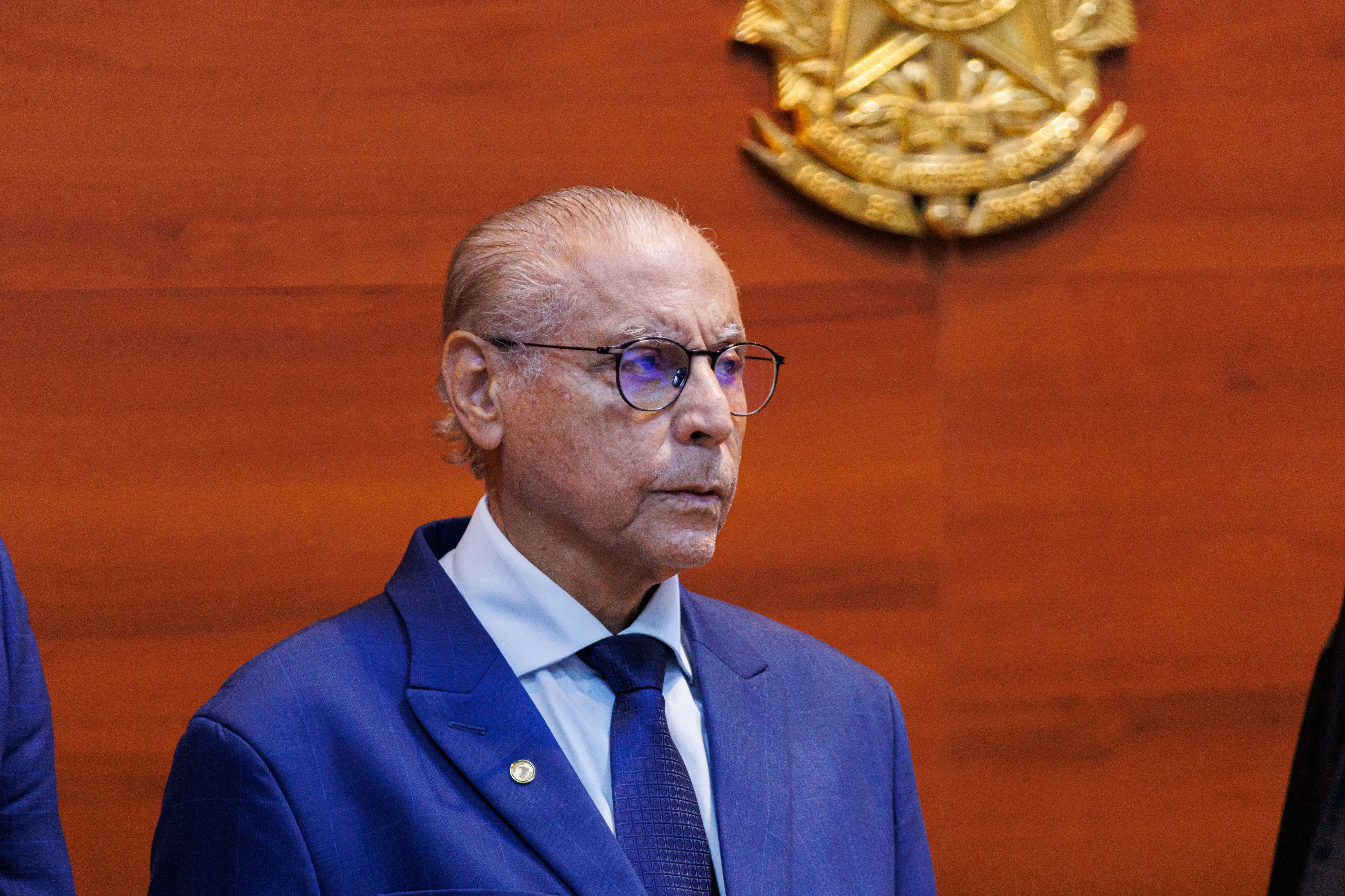 Um homem idoso com pele escura, cabelo branco e óculos está em foco. Ele veste um terno azul e uma gravata azul-escura. Ao fundo, uma parede de madeira com um emblema dourado. Ele tem uma expressão séria e está olhando para a direita.