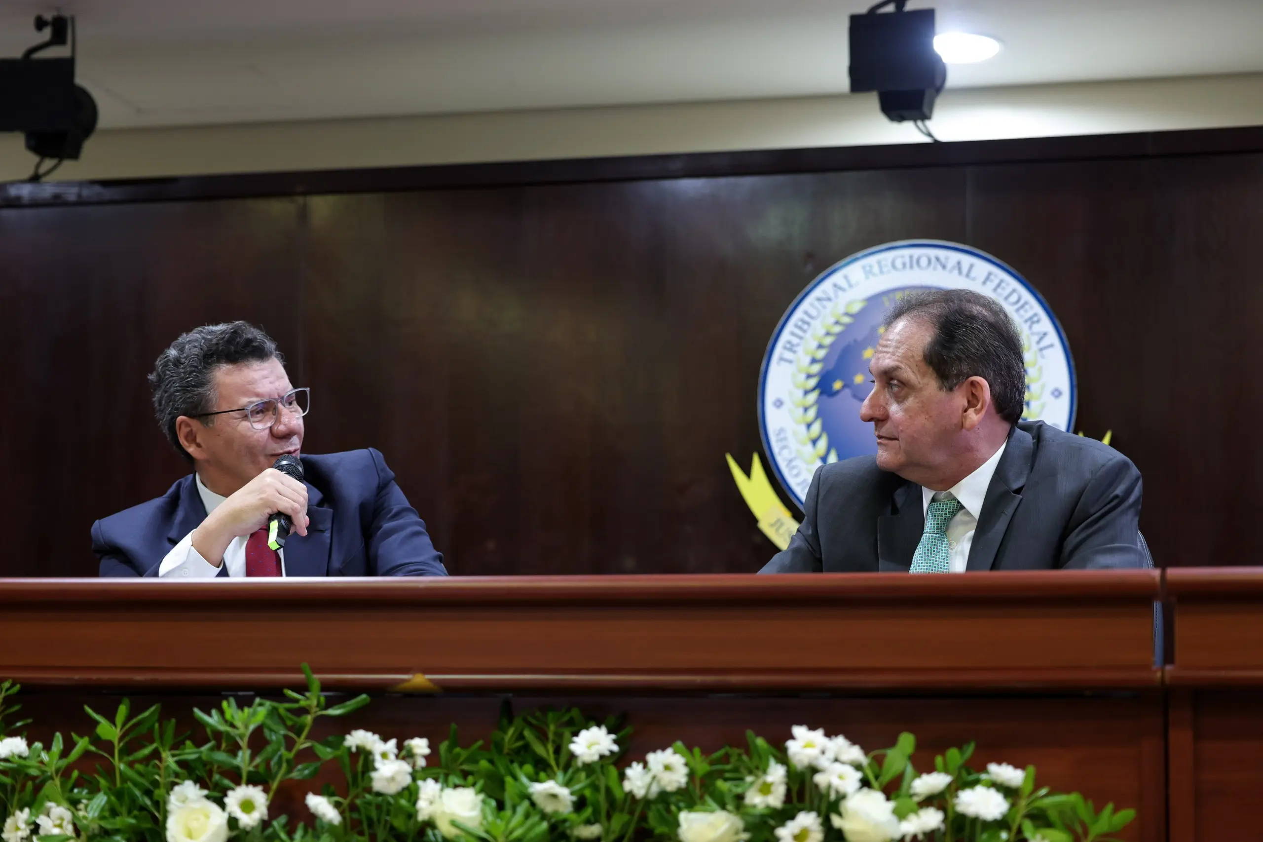 Dois homens de terno sentados atrás de uma bancada decorada com flores brancas na parte inferior. O homem à esquerda fala ao microfone direcionando o olhar ao homem da direita, que o escuta. Ao fundo, o brasão do Tribunal Regional Federal está fixado na parede.