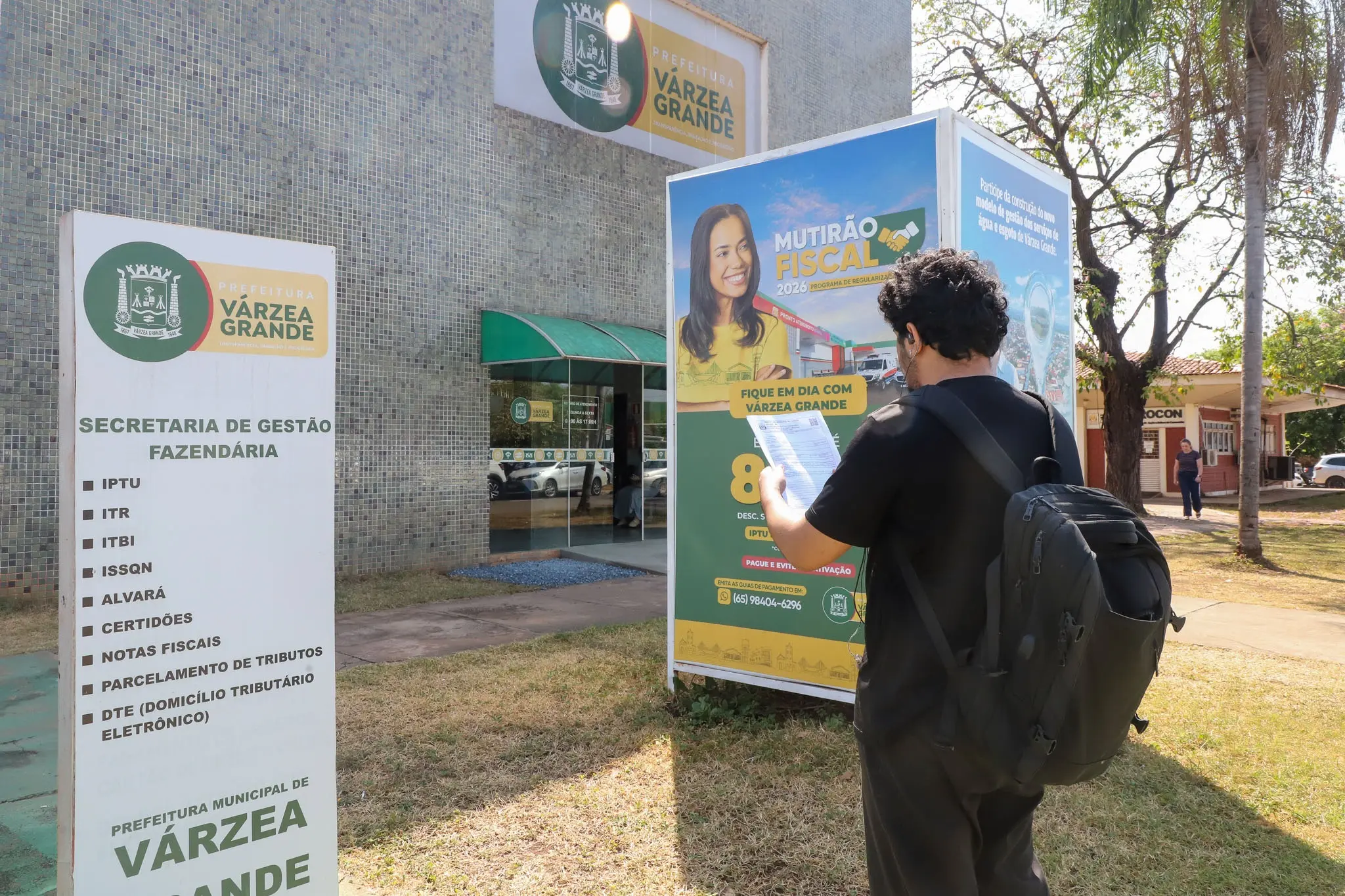Homem consulta material do Mutirão Fiscal 2026 em frente à Secretaria de Gestão Fazendária de Várzea Grande. Ao lado, placas informam serviços tributários e condições para regularização de débitos.