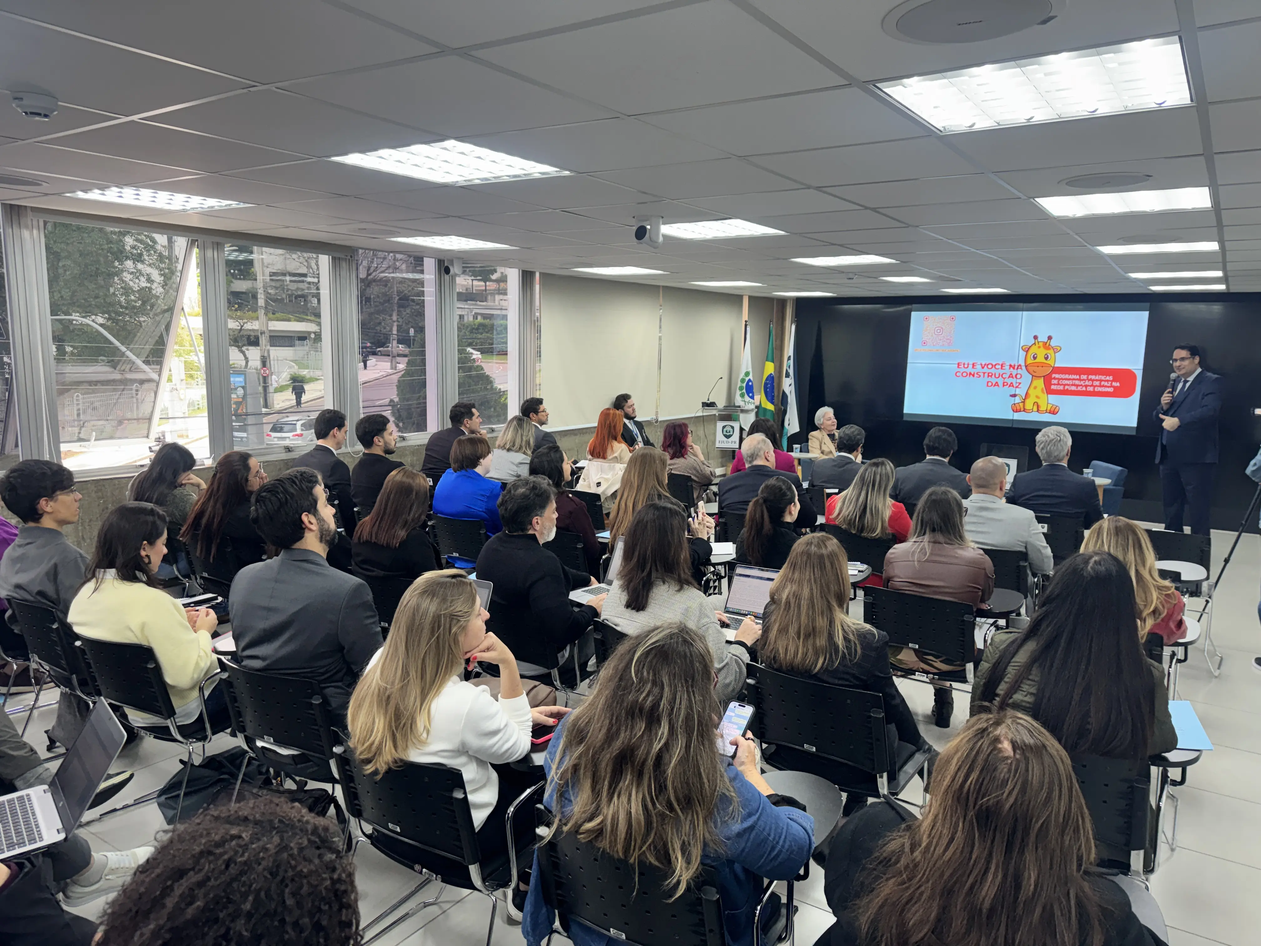 Em imagem horizontal colorida do auditório lotado, o juiz coordenador do Núcleo Gestor da Justiça Restaurativa Túlio Duailibi faz a abertura do painel de lançamento do Sistema Restaura durante o Encontro Paranaense de Justiça Restaurativa. No painel de exposição a logomarca do Programa Eu e Você na